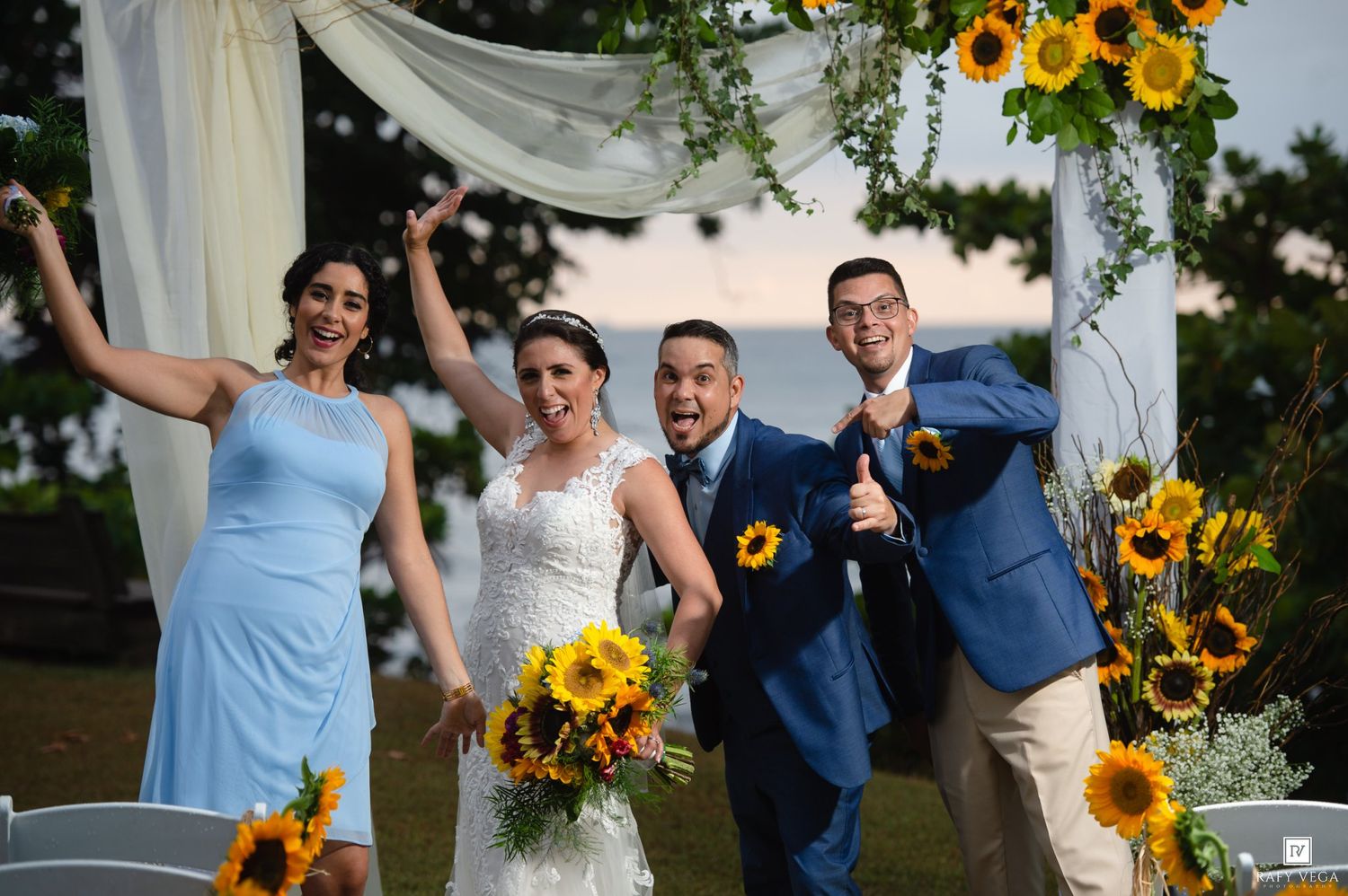 Joyful wedding party celebrates outdoors under sunflower-adorned arch, bride in lace gown with colorful bouquet.