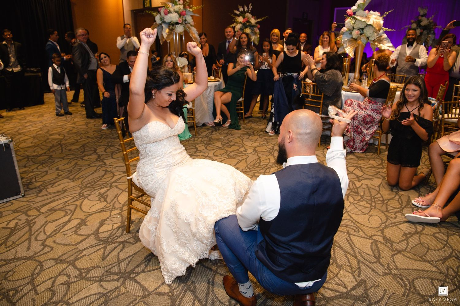 Bride and groom celebrate garter removal tradition at lively indoor wedding reception.