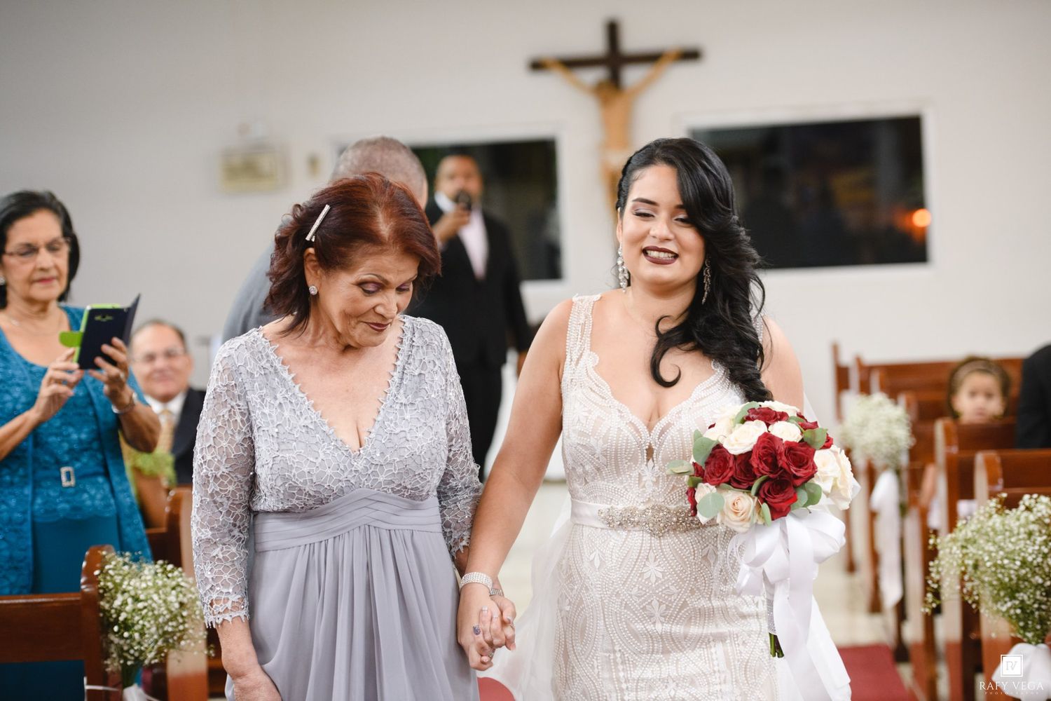 Bride in white lace gown holding red bouquet walks down church aisle with older woman in silver dress.