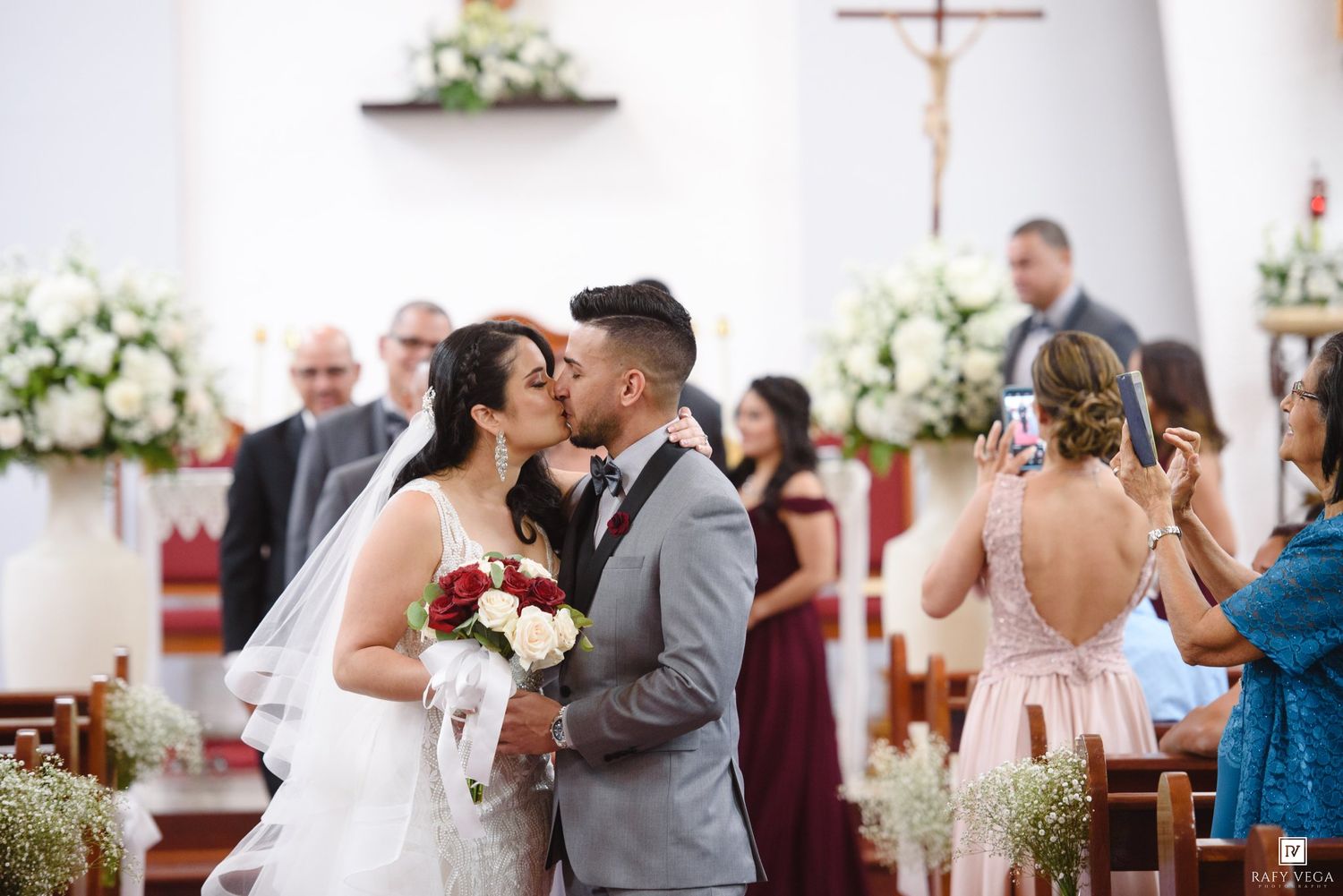 Bride and groom share first kiss at church altar, surrounded by floral arrangements and wedding guests.