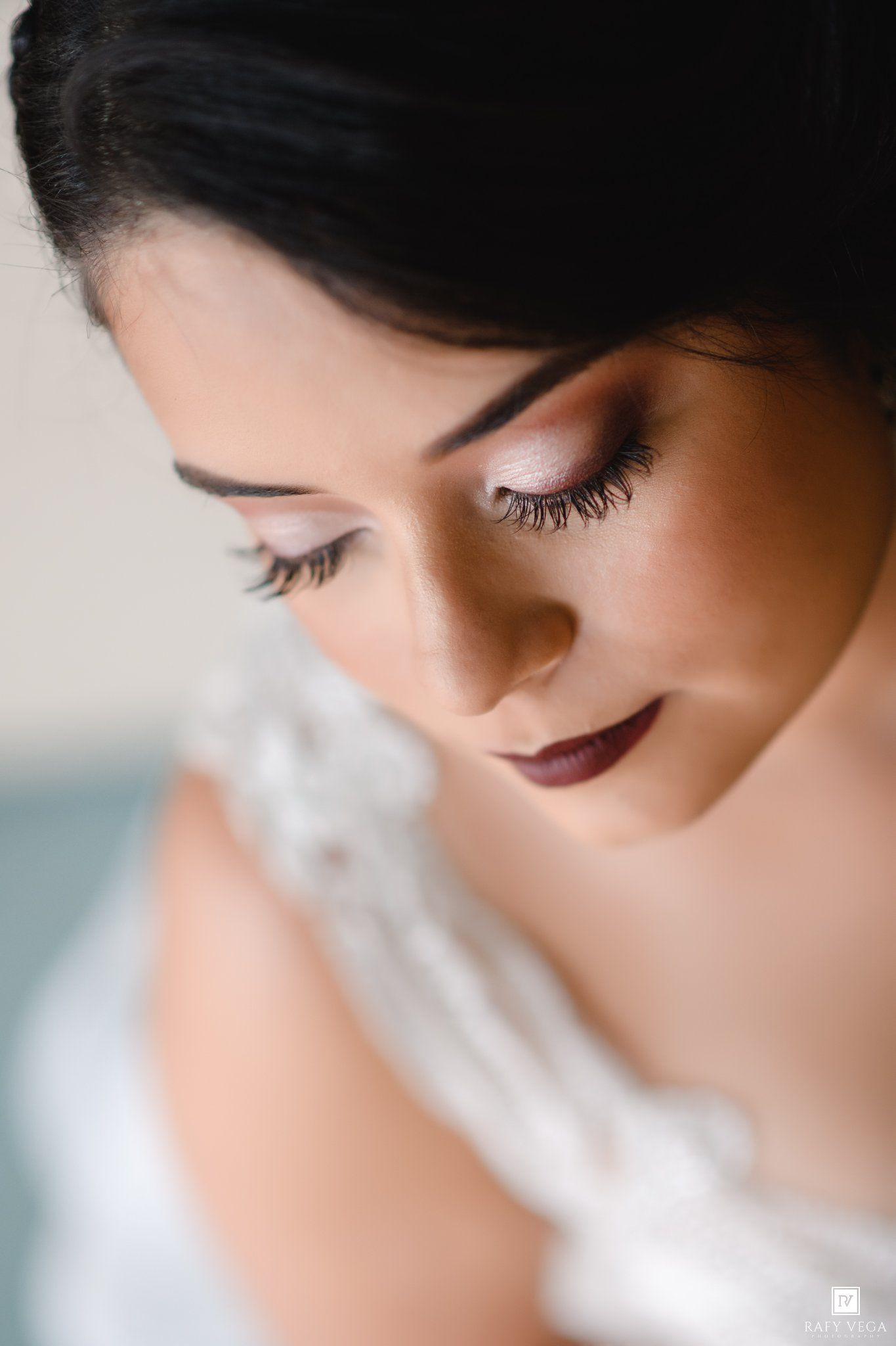 Close-up portrait of a bride with dark hair, smoky eye makeup, and deep berry lips in a white lace wedding dress.