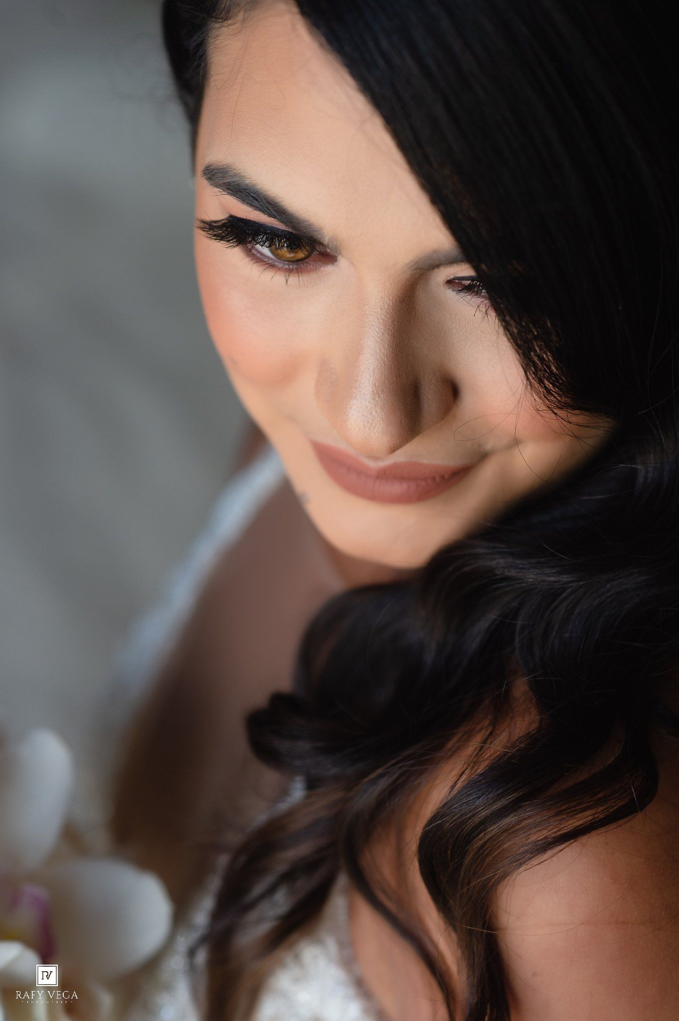 Beautiful bride with dramatic eye makeup and flowing dark hair, smiling softly in her white lace wedding dress.