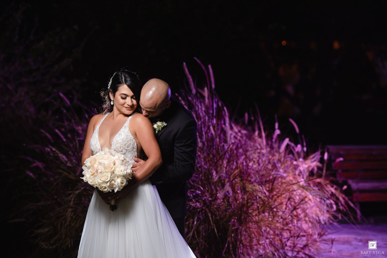 Bride and groom share a romantic embrace outdoors at night with purple-lit ornamental grasses behind them.