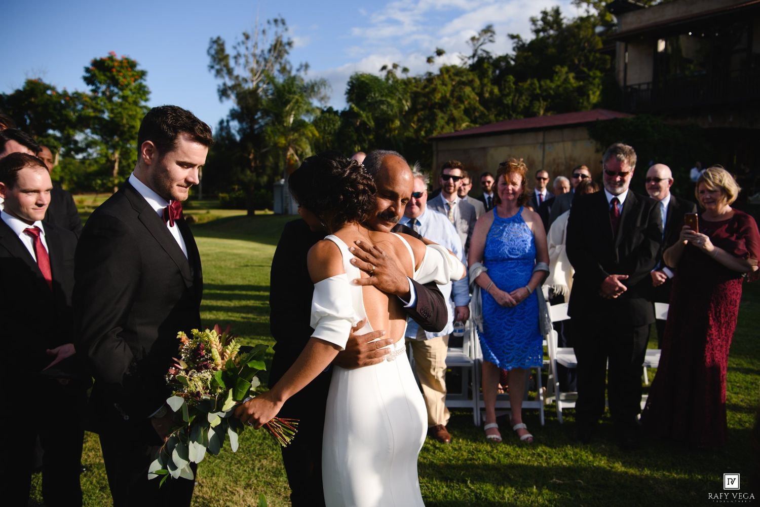 Bride embracing a guest during an outdoor wedding ceremony on a green lawn with attendees watching in the background.