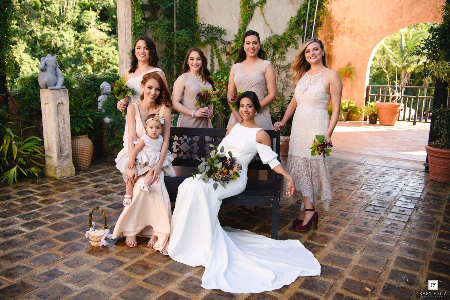 Bride in white gown poses with bridesmaids in champagne dresses and a child in a courtyard garden setting.