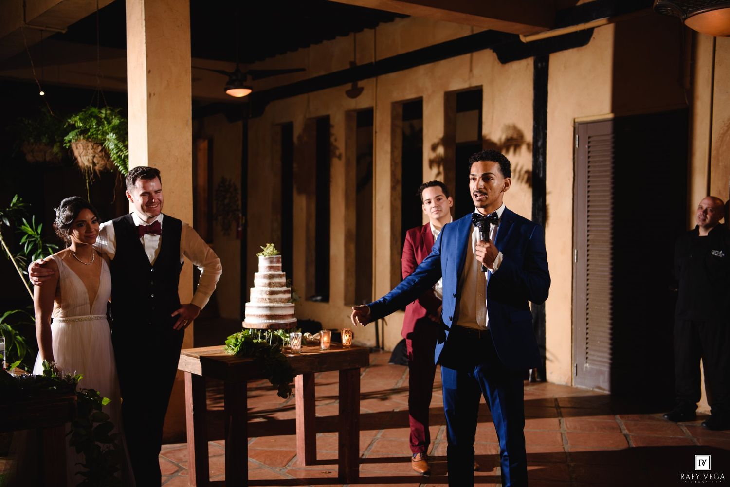 A man in a blue suit gives a speech at a wedding reception while the bride and groom stand near a tiered cake.