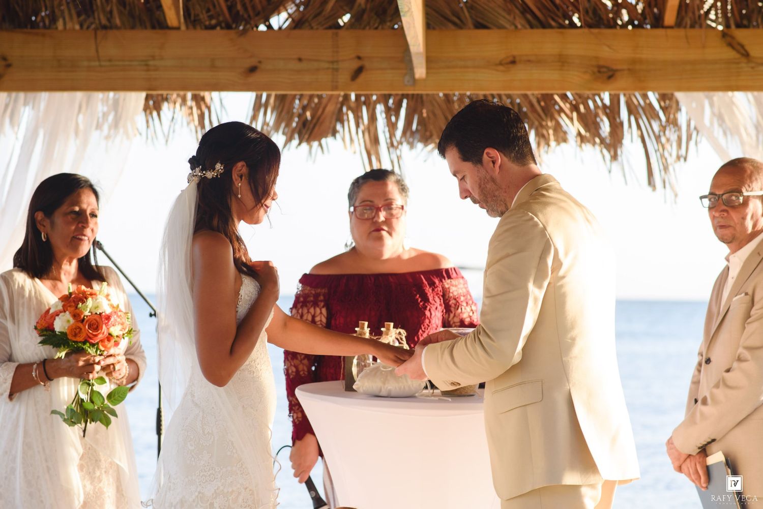 Bride and groom exchange rings during an outdoor beach wedding ceremony under a thatched palapa roof.