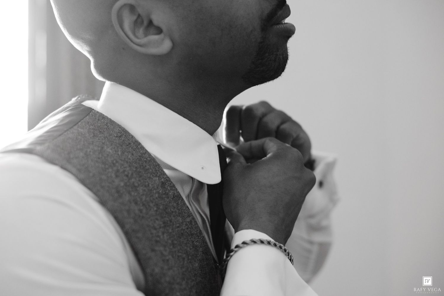 Black and white photo of a groom adjusting his tie while wearing a vest and bracelet on his wedding day.