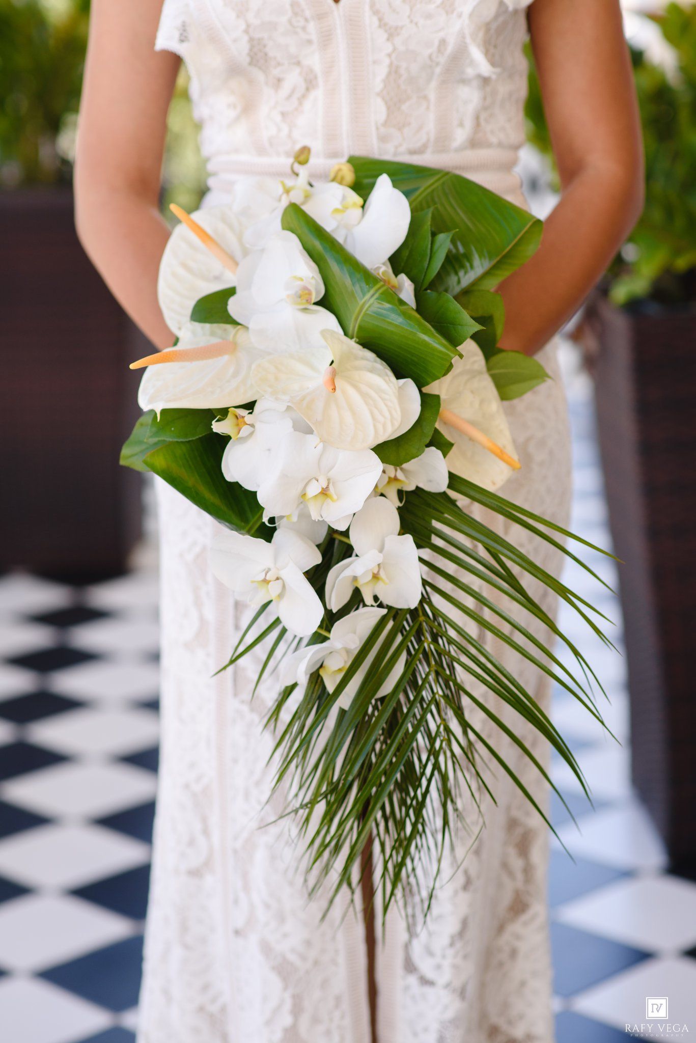Bride holding a tropical wedding bouquet with white orchids, anthuriums, and lush green palm leaves in an elegant cascade style.