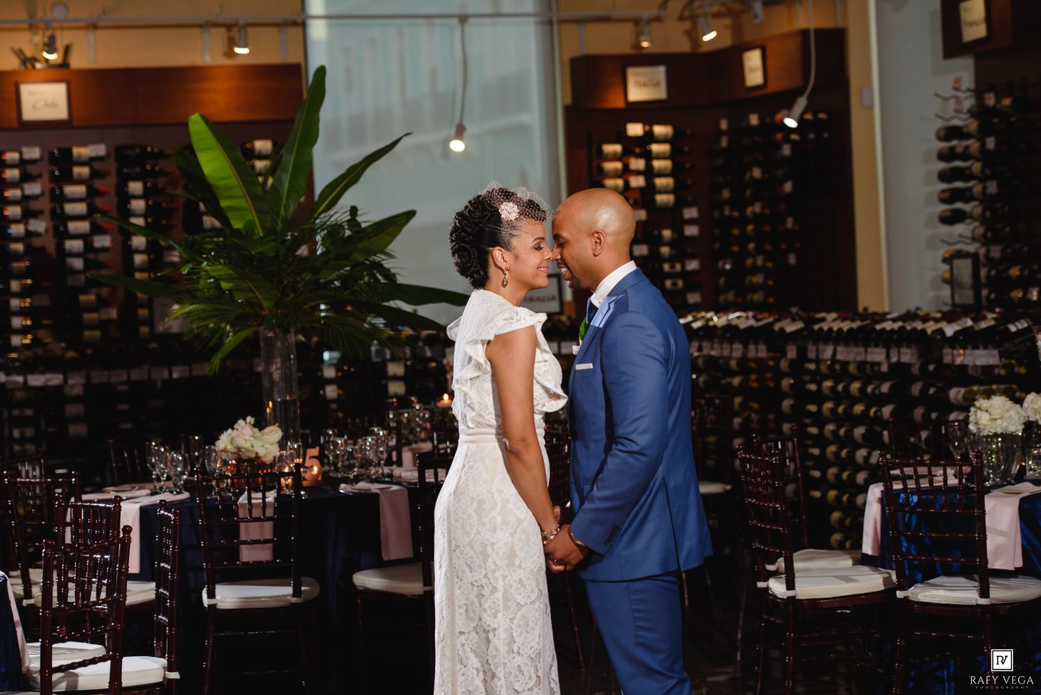 A couple shares a romantic moment in a wine bar wedding venue, the bride in lace and groom in a blue suit.