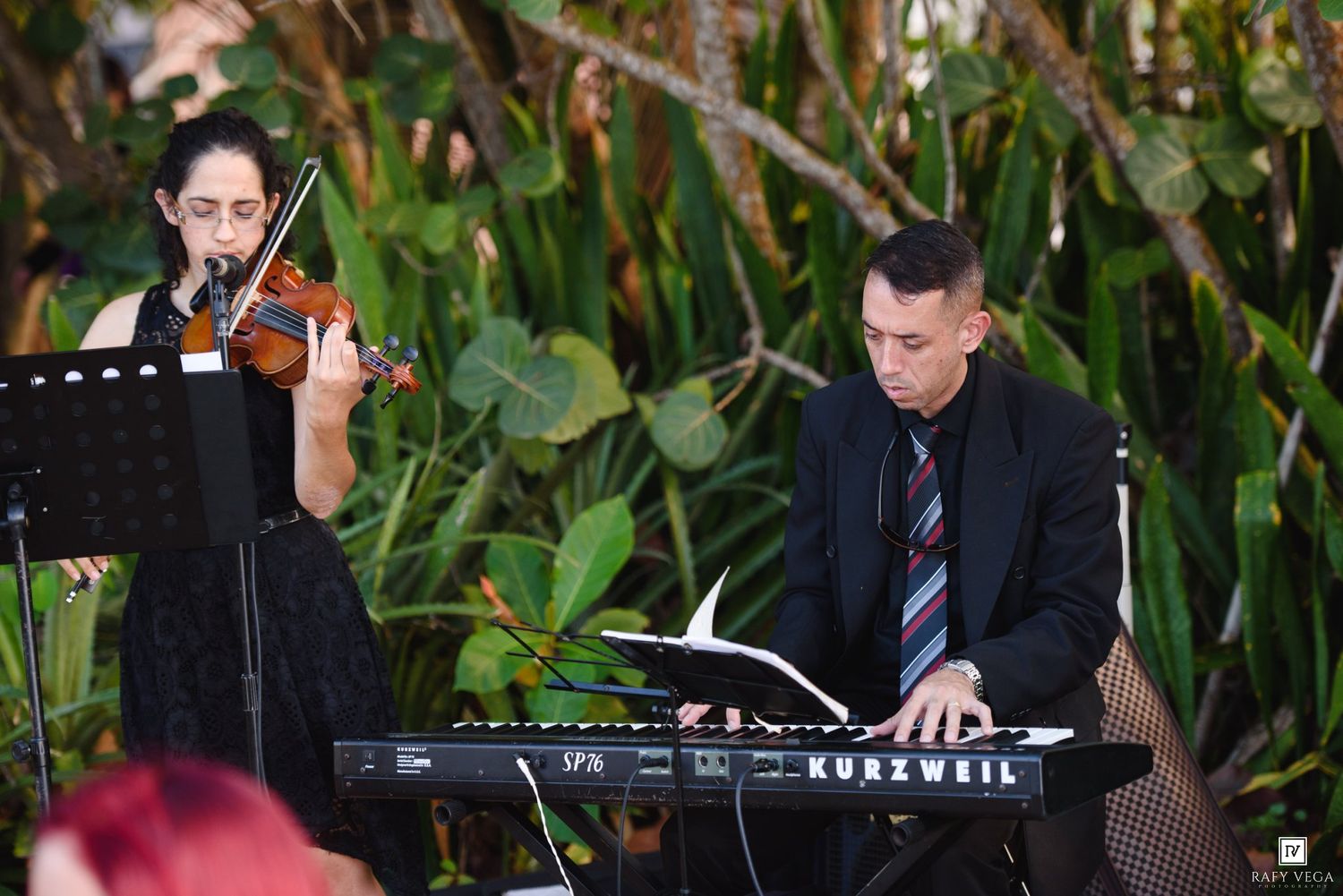 Live wedding musicians perform with violin and Kurzweil keyboard amid lush tropical greenery.