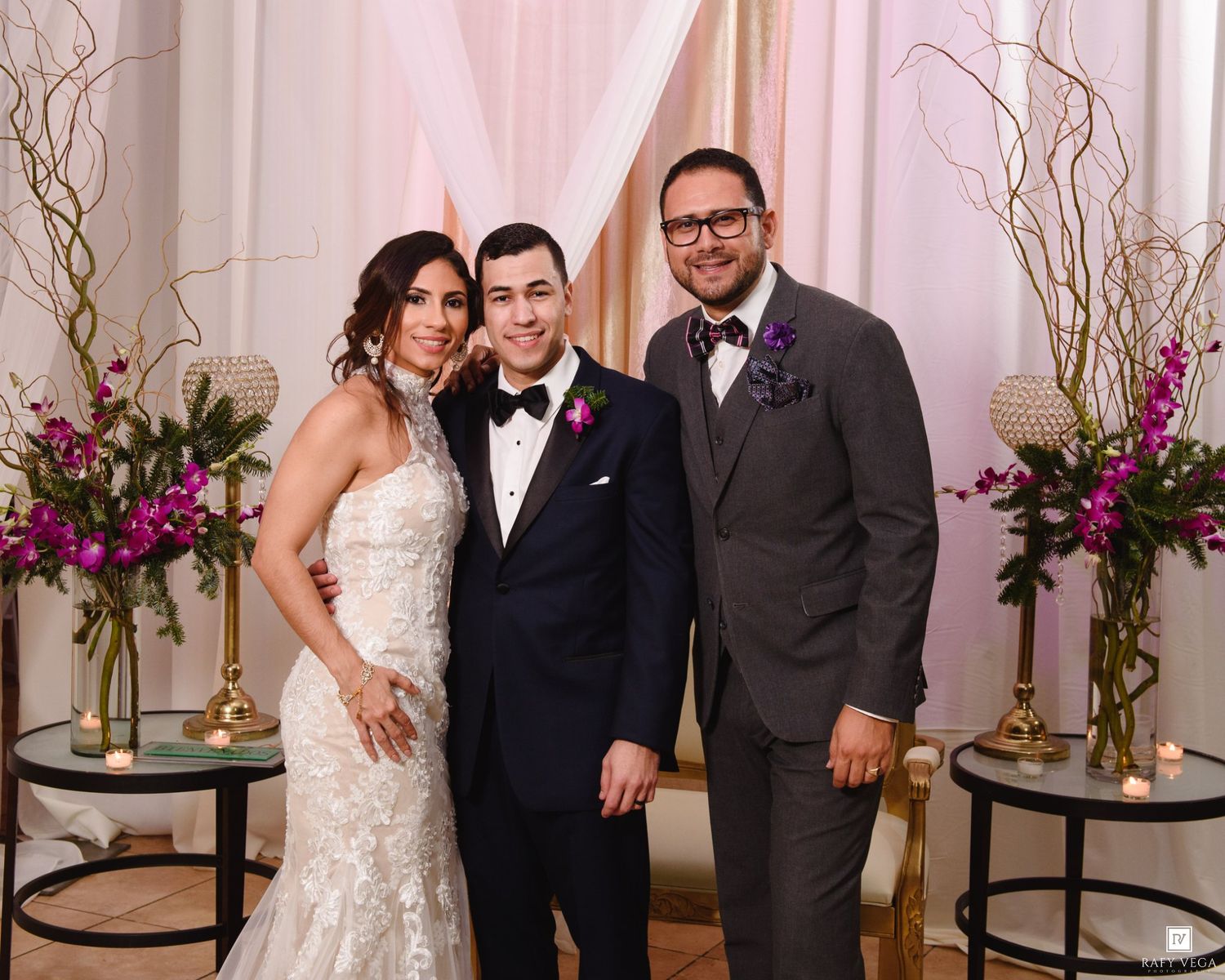 Bride in lace gown and groom in tuxedo pose with guest at elegant wedding ceremony with floral decor.