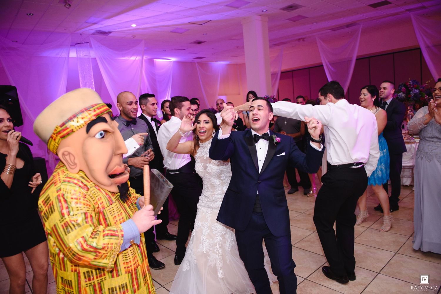 Bride and groom dancing joyfully with a costumed character at a wedding reception with purple lighting.