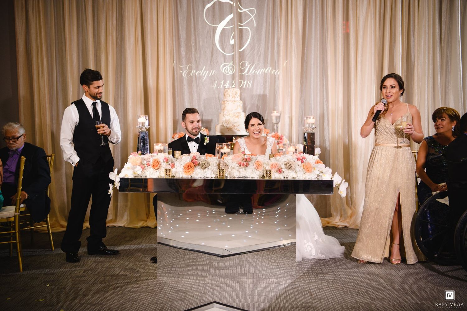 Wedding reception toast with bride and groom seated at sweetheart table while bridesmaid speaks into microphone.