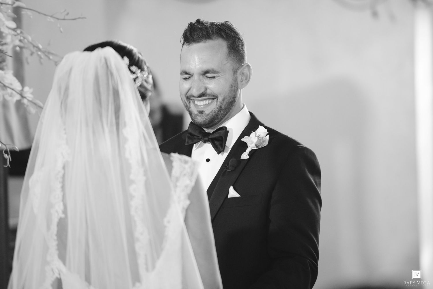 Black and white photo of a smiling groom in a tuxedo facing his veiled bride during a wedding ceremony.