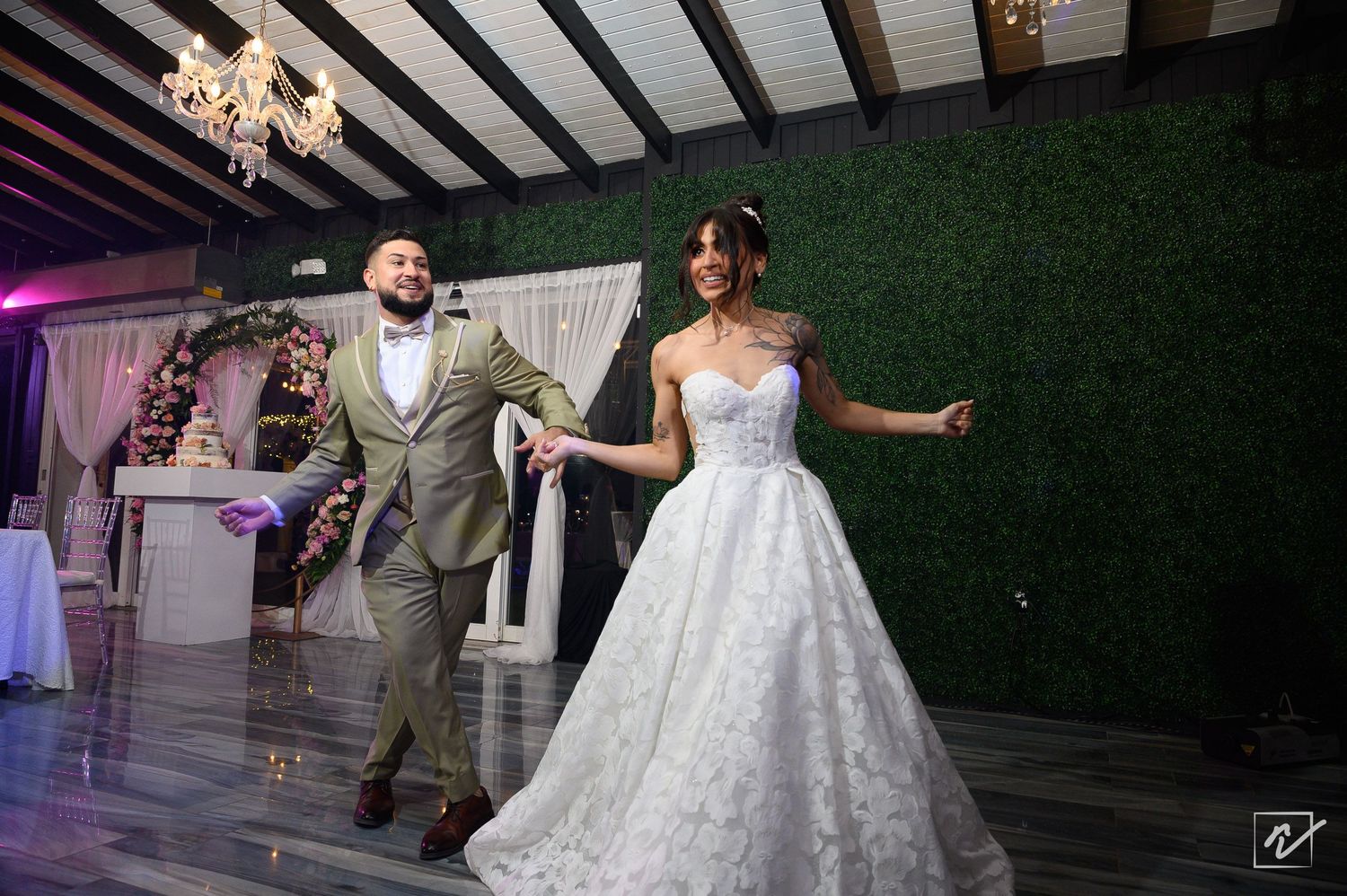 Smiling bride in a white ball gown holds hands with groom in a tan suit as they dance at an elegant wedding reception with chandeliers.