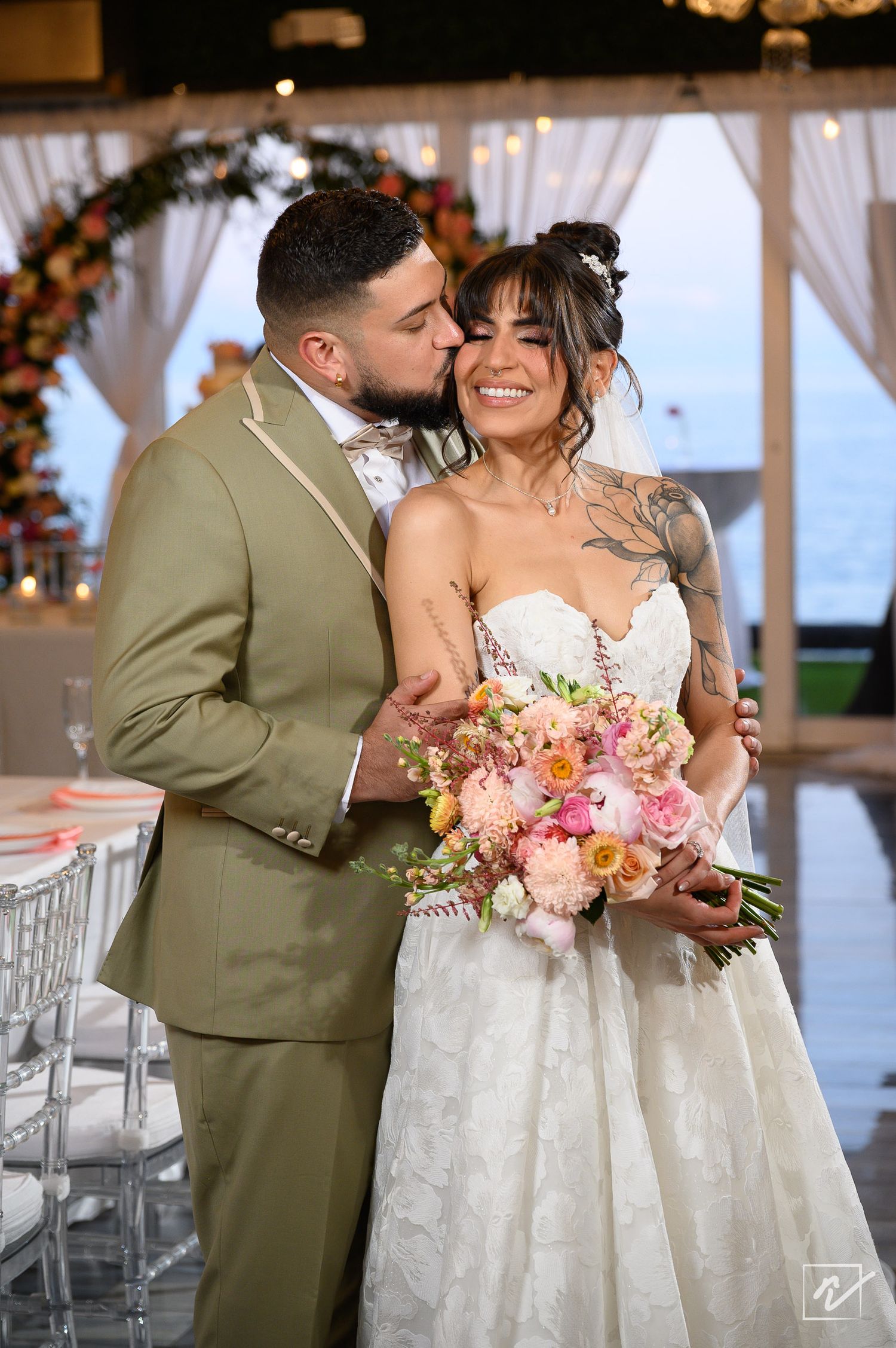 Smiling bride in a white lace gown holding a pink floral bouquet is kissed on the cheek by her groom in an olive suit at a wedding venue.