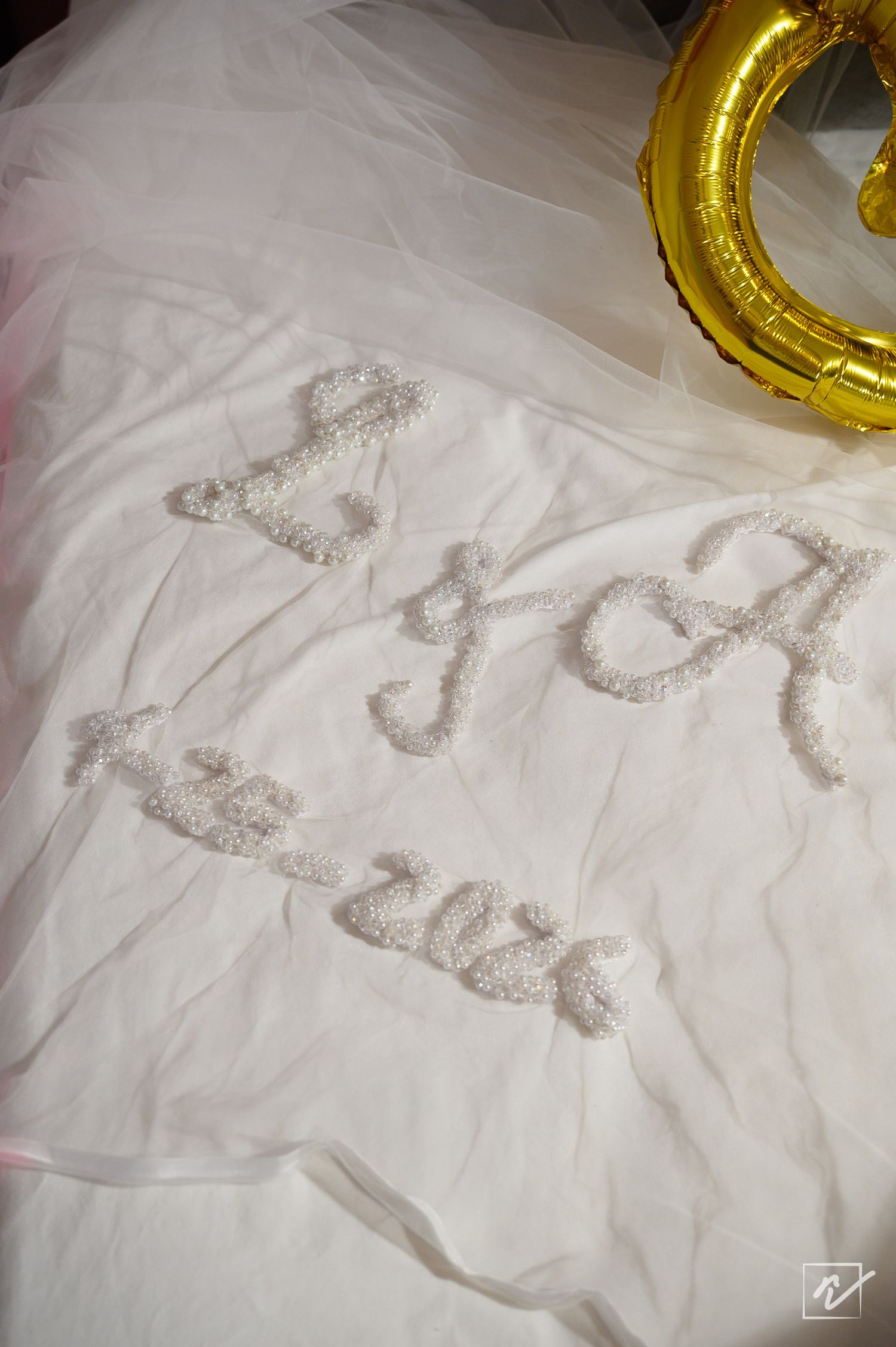 Wedding veil with hand-beaded white initials and date embroidered on tulle, beside a gold ring-shaped balloon.