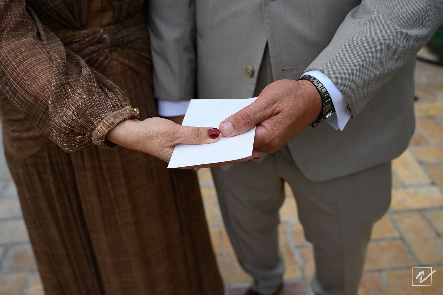 Close-up of two people standing together, one in a brown plaid dress and one in a gray suit, holding a folded white note in their hands.