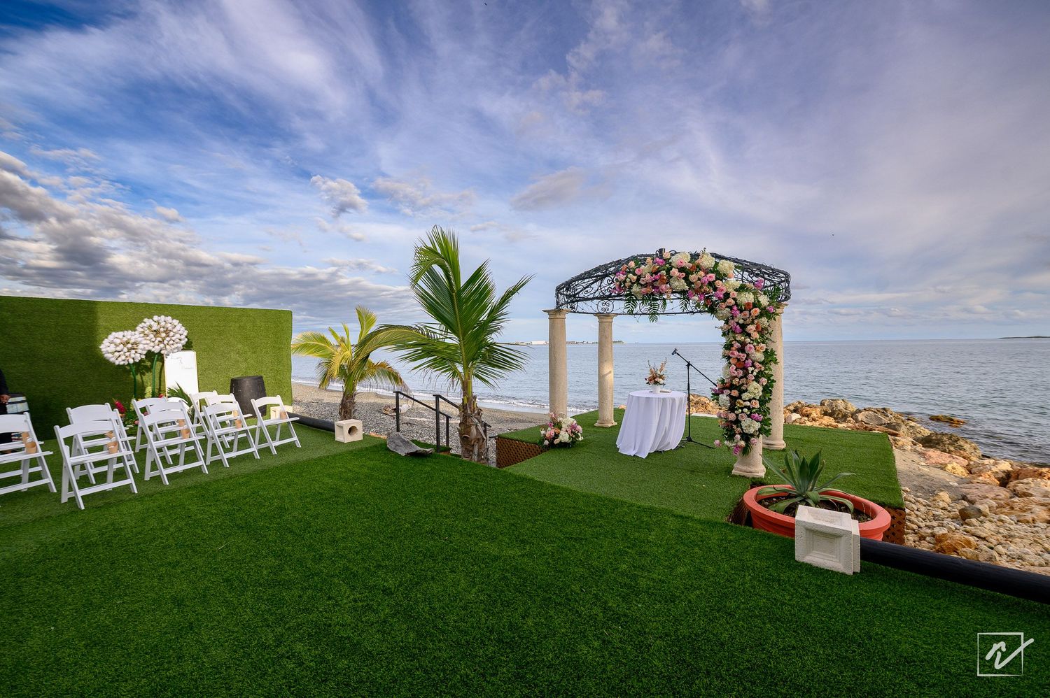 Oceanfront wedding ceremony setup with a floral arch, white chairs, and palm trees on a green lawn overlooking the sea at sunset.