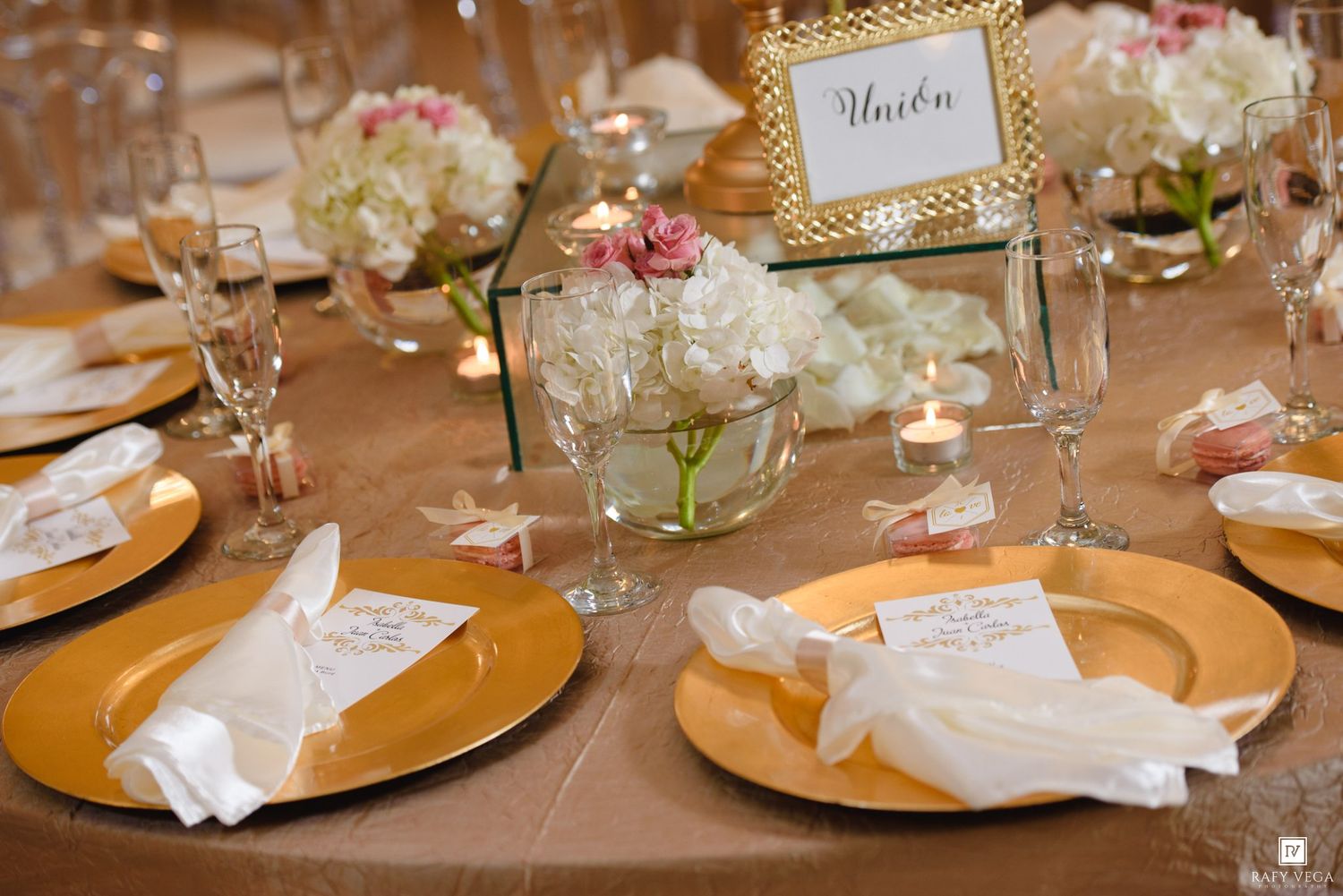 Elegant wedding reception table with gold charger plates, white napkins, floral centerpieces, and glowing candles.