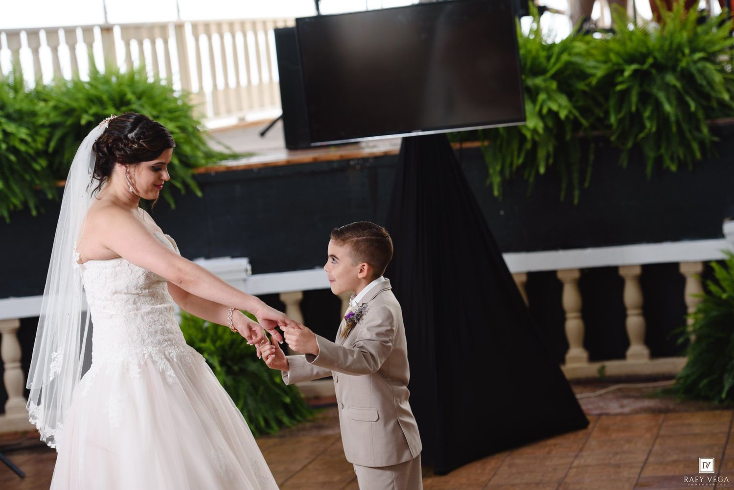 Bride in white wedding gown dancing with young boy in tan suit at outdoor wedding reception.