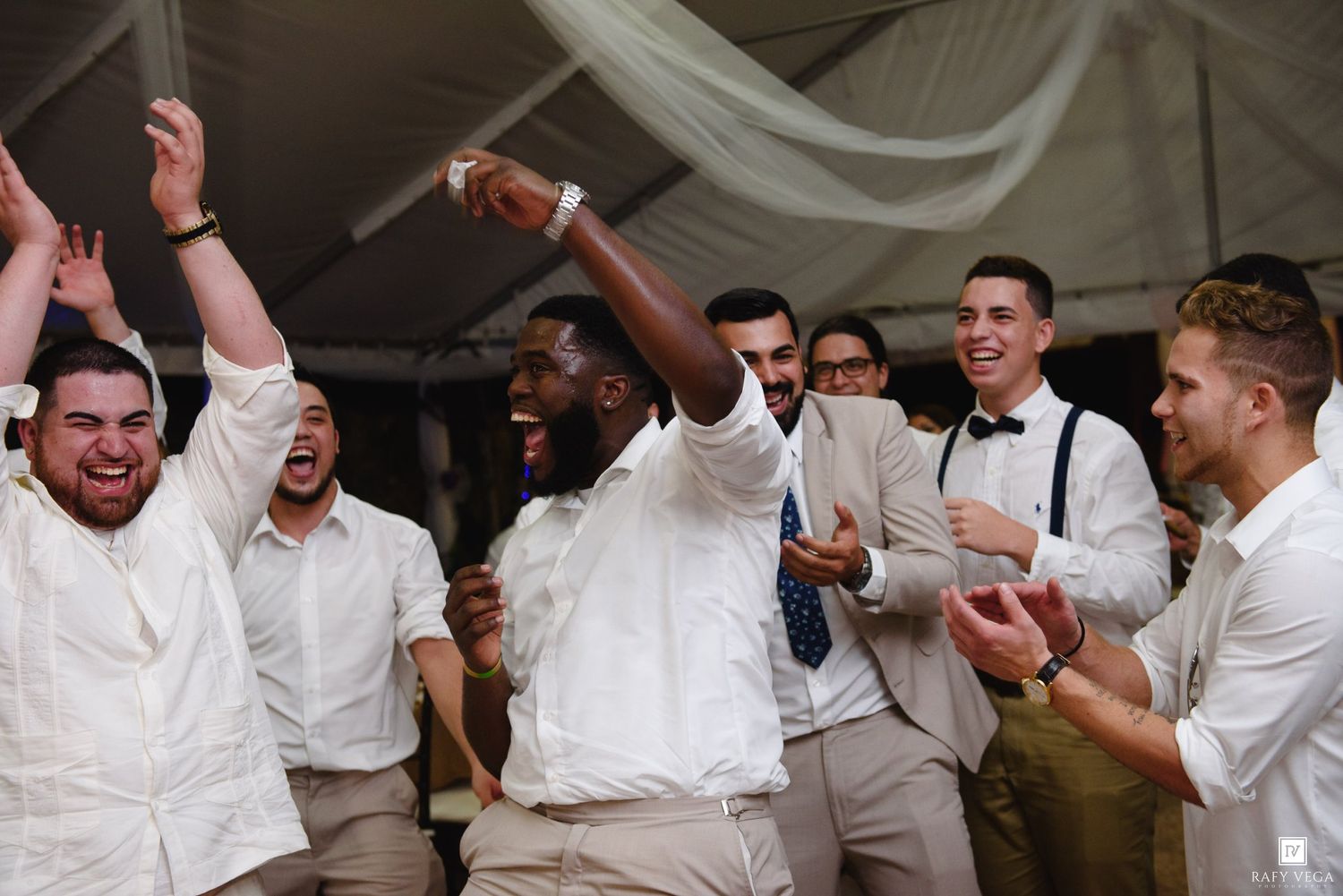 Groomsmen celebrating joyfully at a wedding reception, wearing white shirts and khaki pants under a tent.