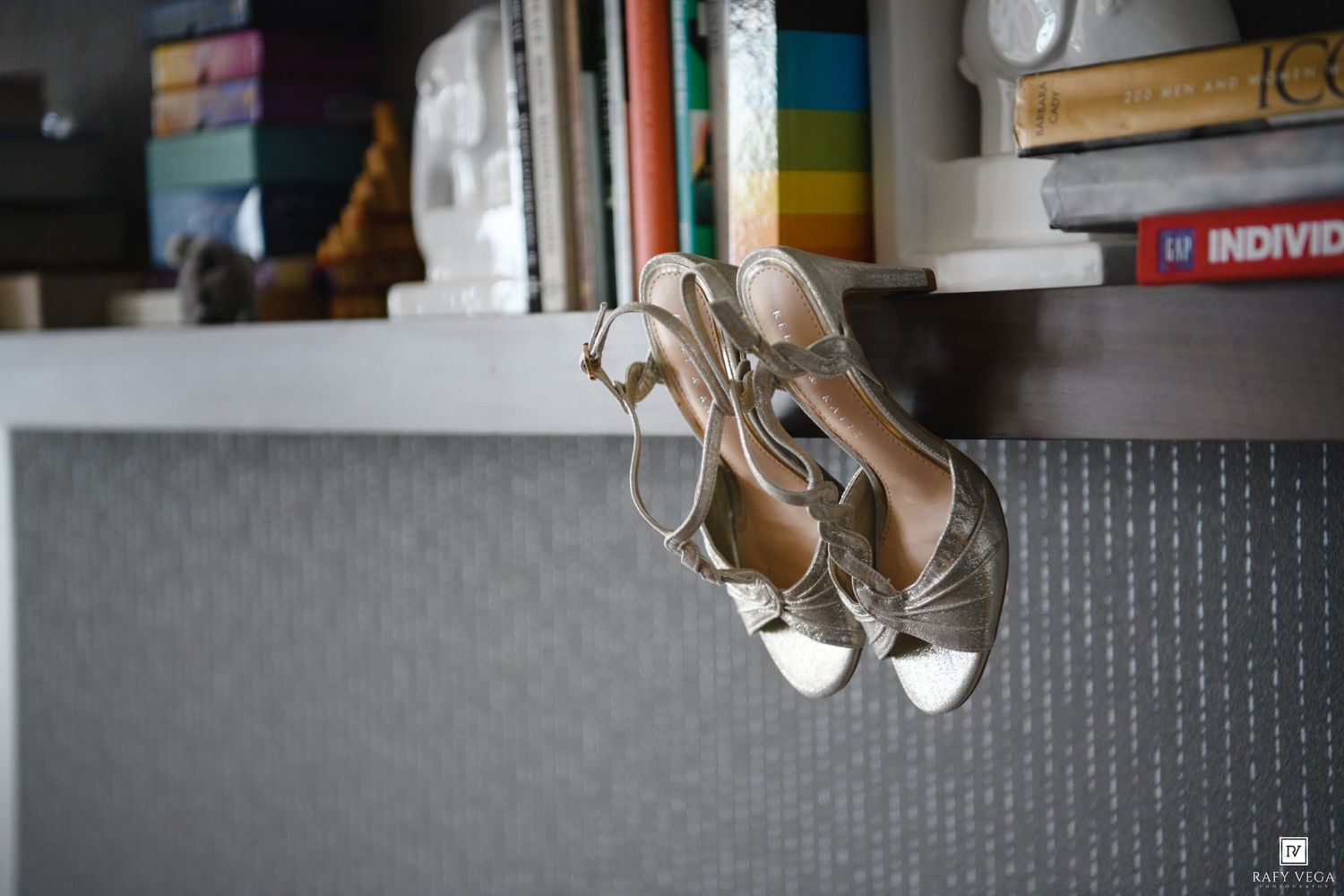 Gold strappy high heel sandals hanging on a shelf beside colorful books and decorative items.