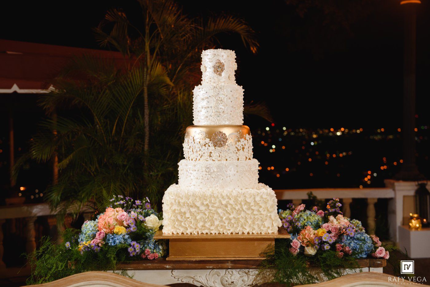 Elegant five-tier white wedding cake with gold accents displayed outdoors at night with colorful floral arrangements.
