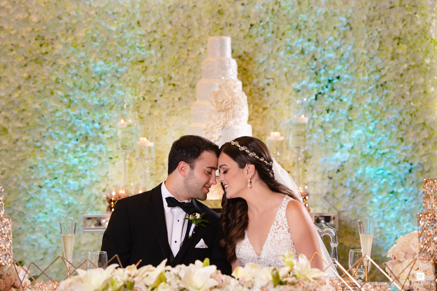 Bride and groom share a romantic forehead touch at sweetheart table with floral wall and wedding cake backdrop.