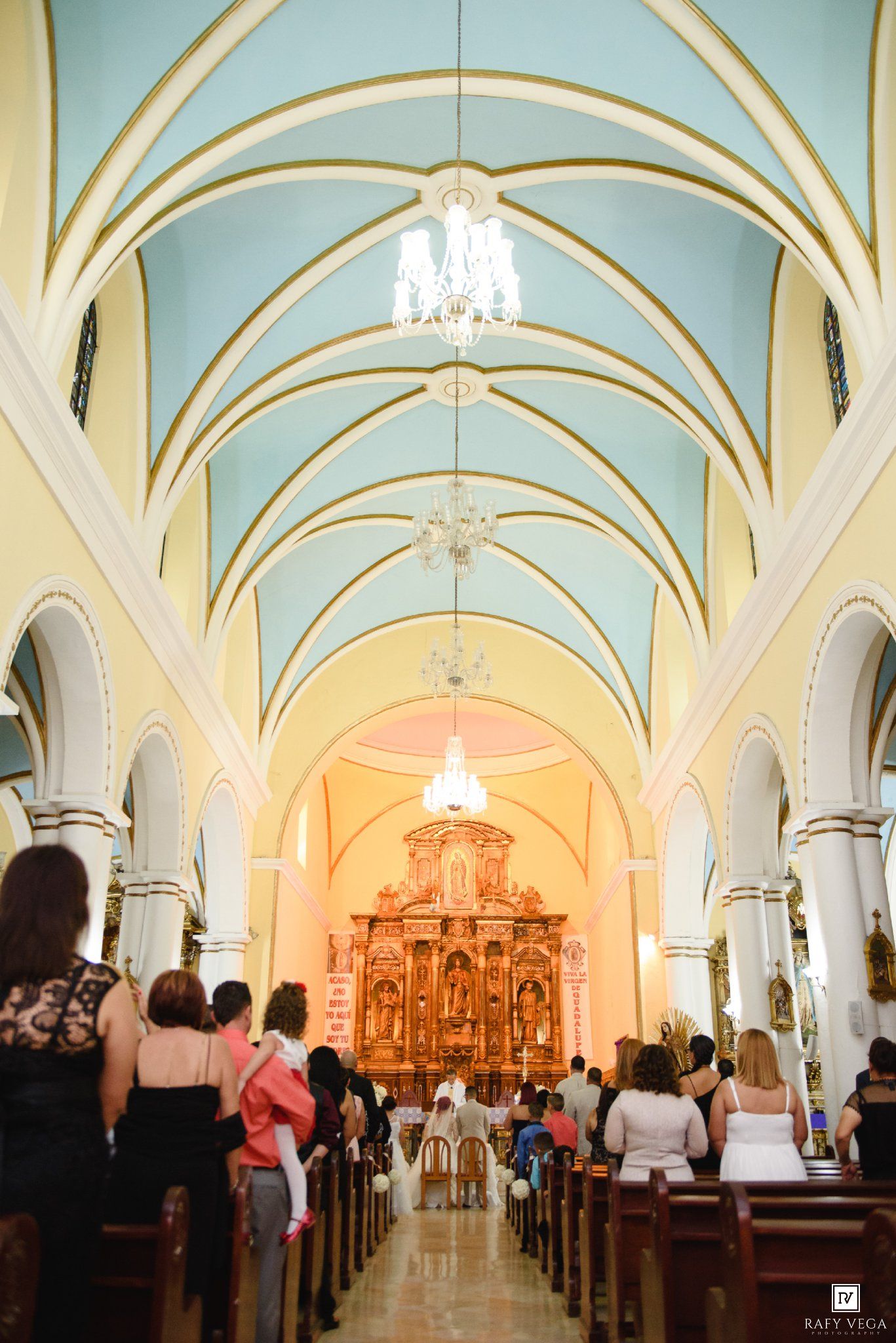 Wedding ceremony inside ornate church with blue vaulted ceiling and golden altar in background.