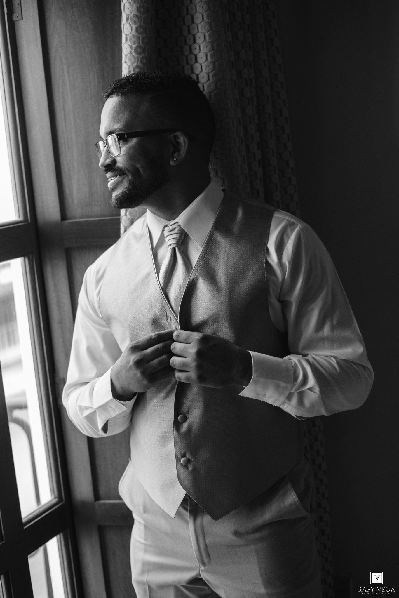 Black and white photo of a groom adjusting his vest and tie by a window in formal wedding attire.