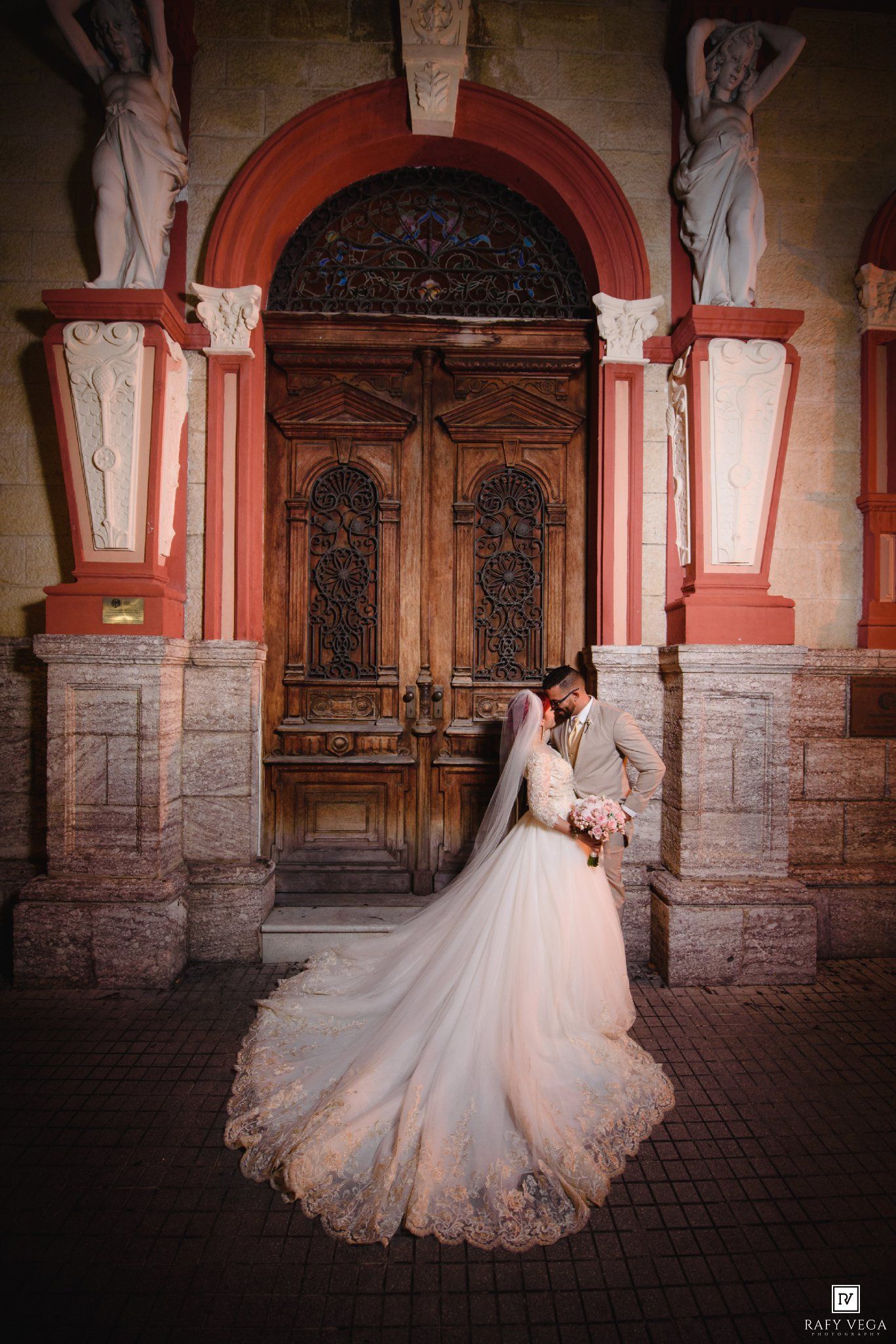 Bride and groom embracing before ornate wooden church doors with classical statues and pink columns.