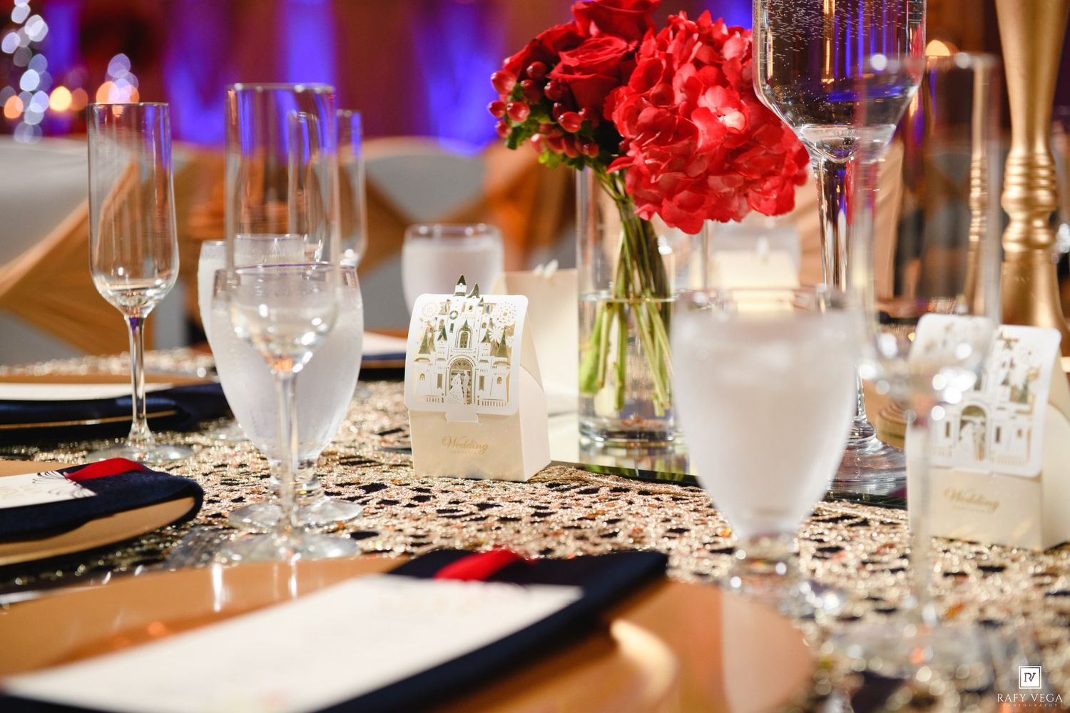 Elegant wedding reception table setting with red floral centerpiece, crystal glassware, and gold sequin tablecloth.