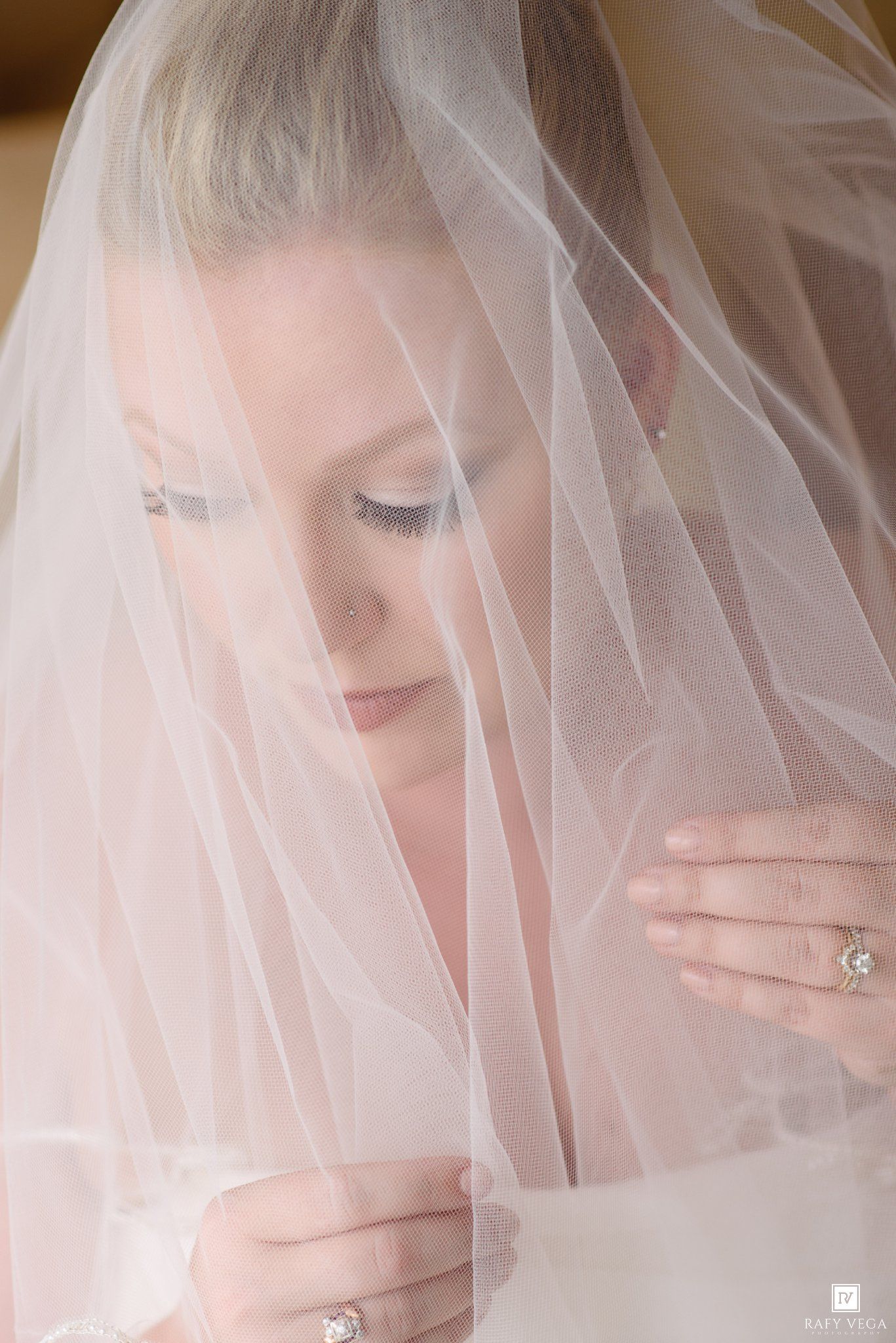 A bride gazes down under her white veil, showcasing her diamond engagement ring and elegant bridal makeup.