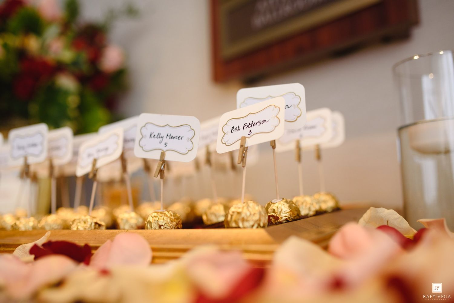 Wedding escort card display with Ferrero Rocher chocolates and rose petals on a wooden tray at an elegant reception.