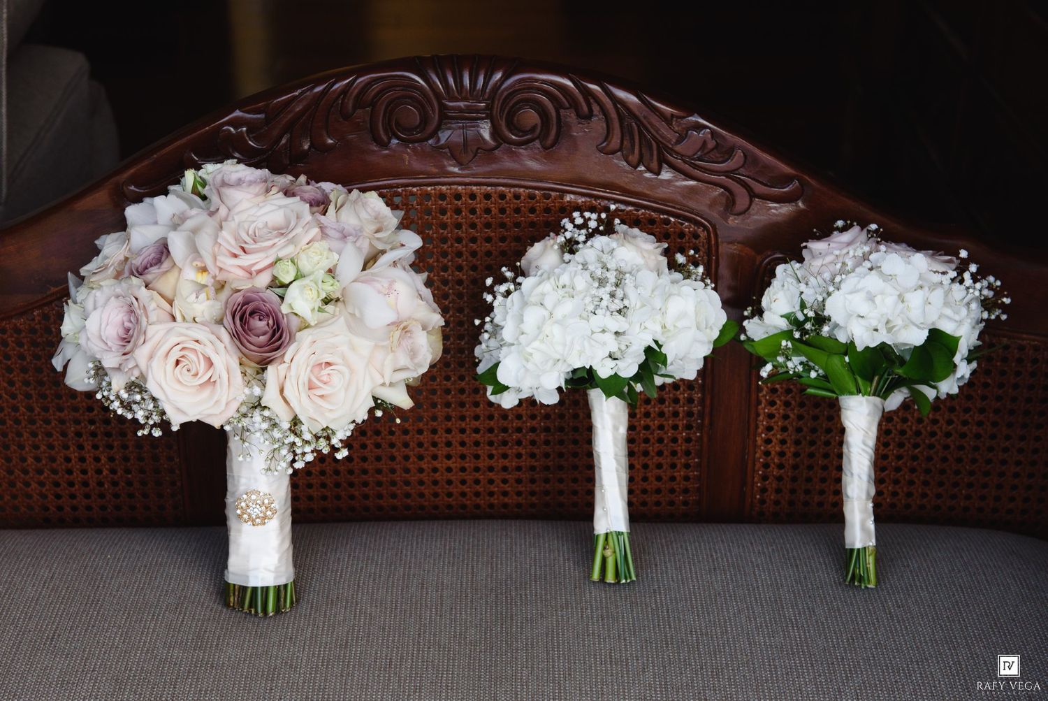 Three elegant wedding bouquets with white hydrangeas, baby's breath, and blush roses rest on a wooden chair.
