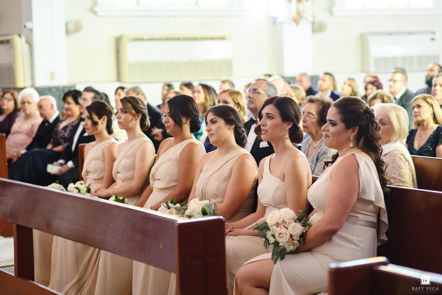 Bridesmaids in blush gowns holding white bouquets seated in church pews during a wedding ceremony.