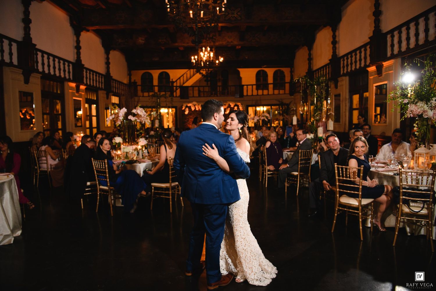 Couple shares first dance at elegant wedding reception in ornate ballroom with golden chairs and warm chandelier lighting.