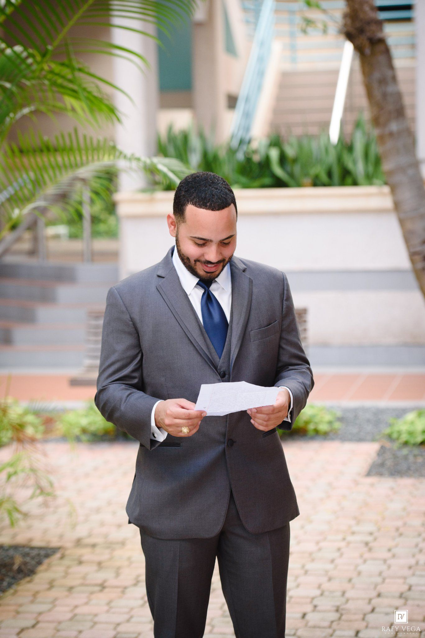 A well-dressed man in a gray suit and blue tie smiles while reading a document outdoors.