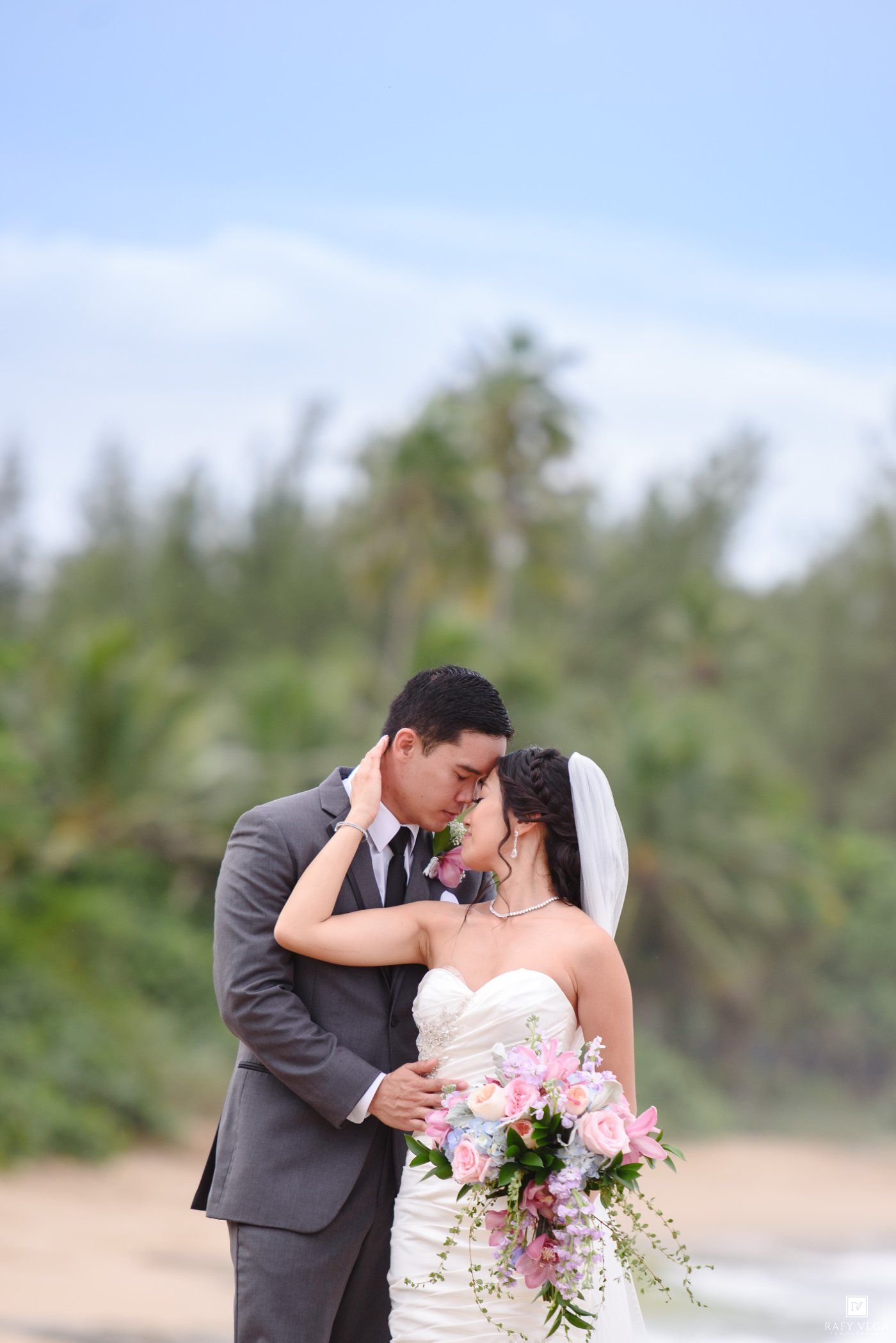 Bride and groom share a romantic moment on a tropical beach, bride holding pink floral bouquet in white gown.