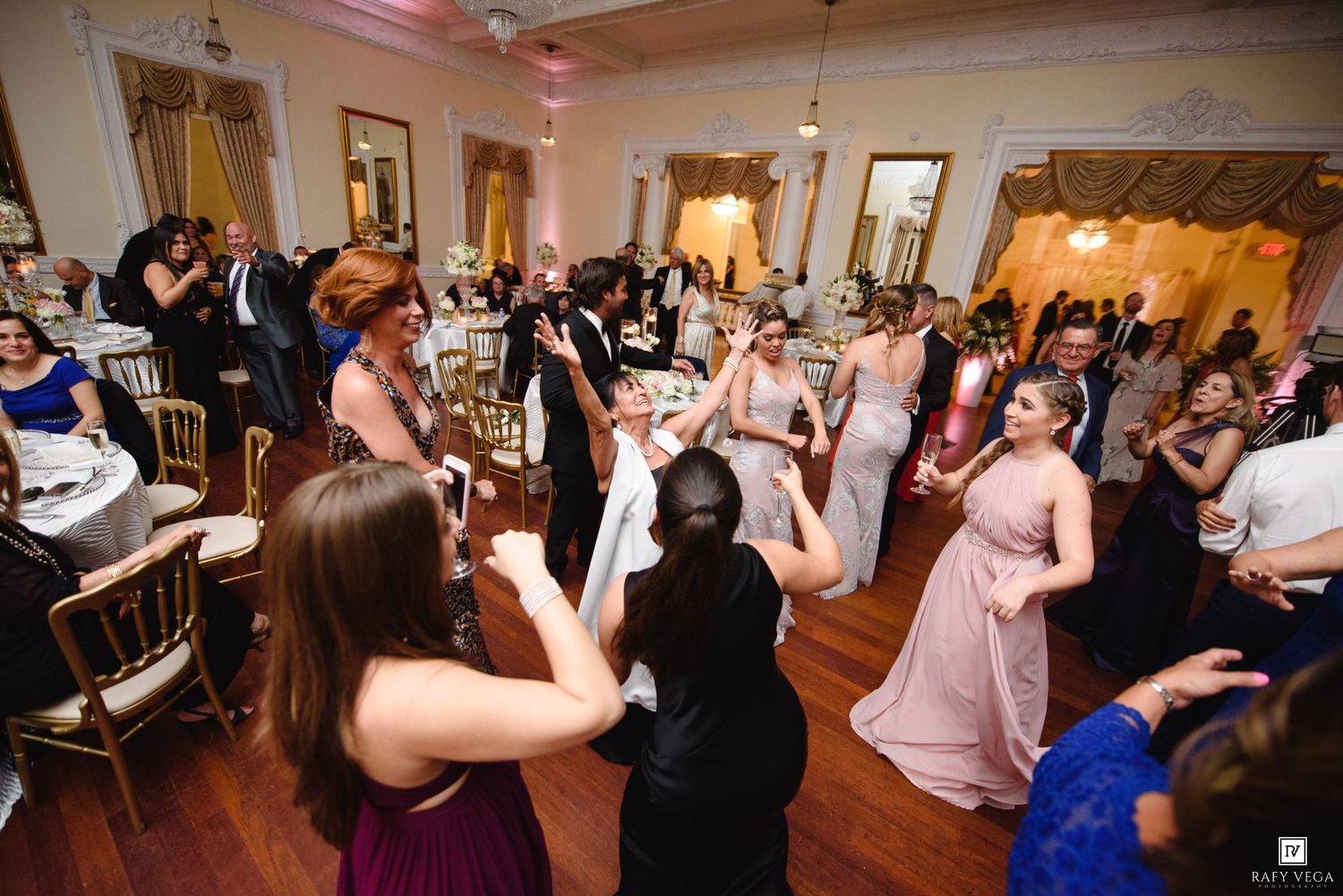 Guests dancing joyfully at an elegant wedding reception in a grand ballroom with chandeliers and formal attire.