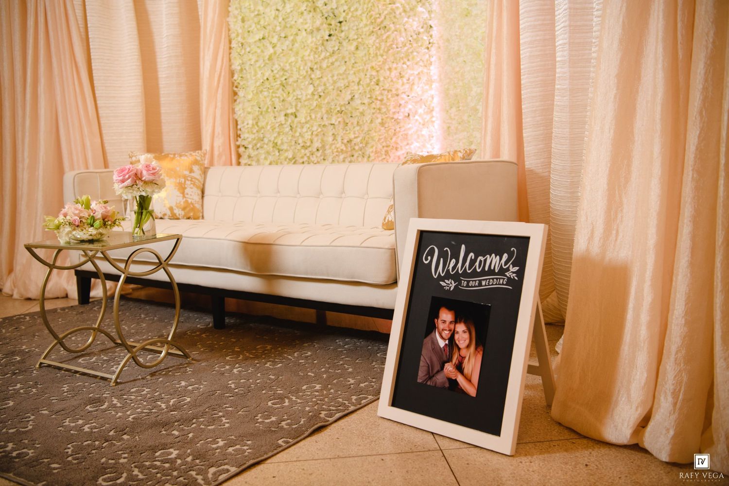 Elegant wedding reception lounge with white sofa, floral wall backdrop, and a welcome sign with couple photo.