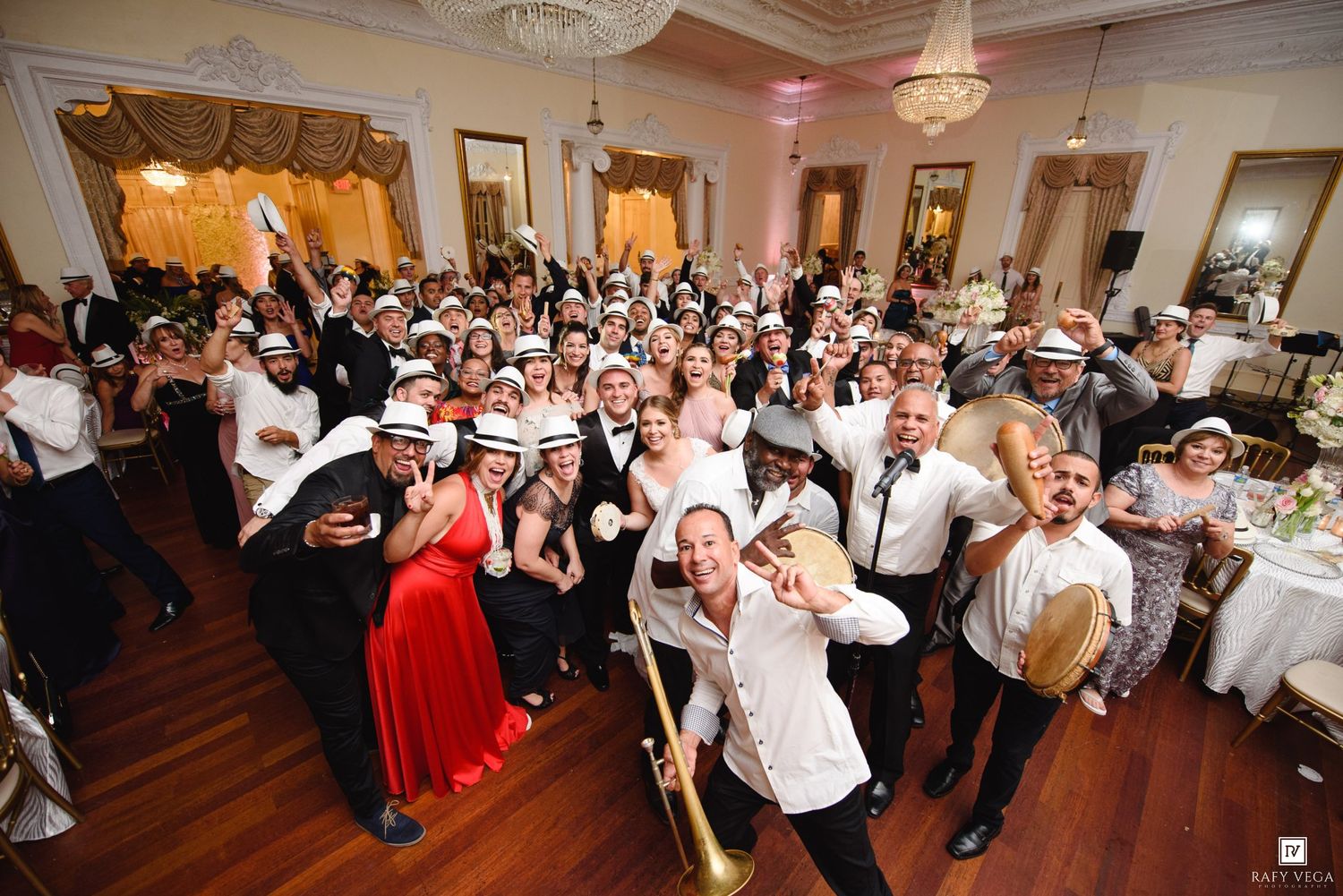 Large wedding reception group photo with guests wearing white fedoras, dancing in elegant ballroom with chandelier.