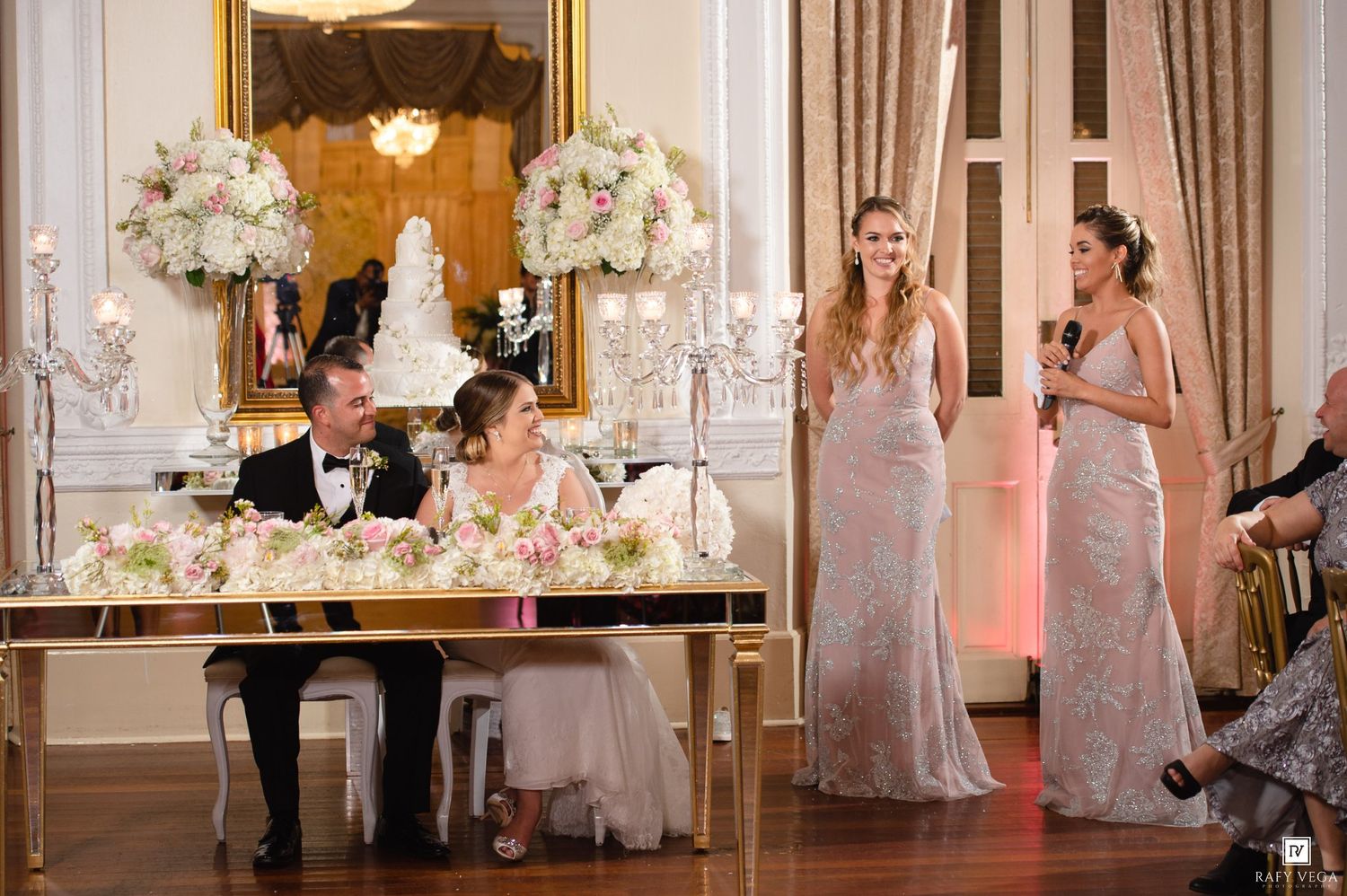 Two bridesmaids in sequin gowns give a toast at an elegant wedding reception with floral arrangements and a white cake.