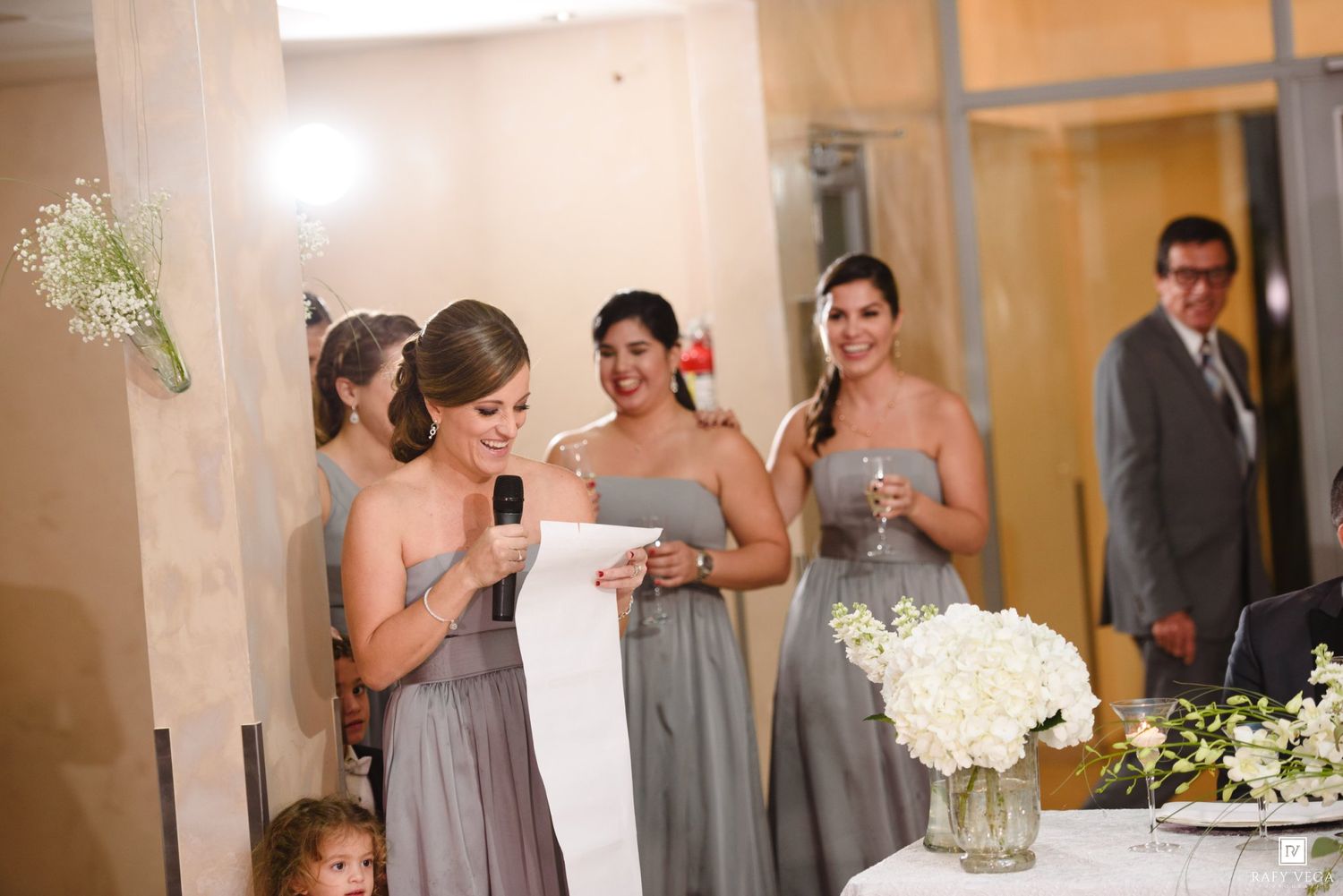 Bridesmaids in gray dresses laughing during wedding reception speech, holding champagne glasses near floral centerpiece.