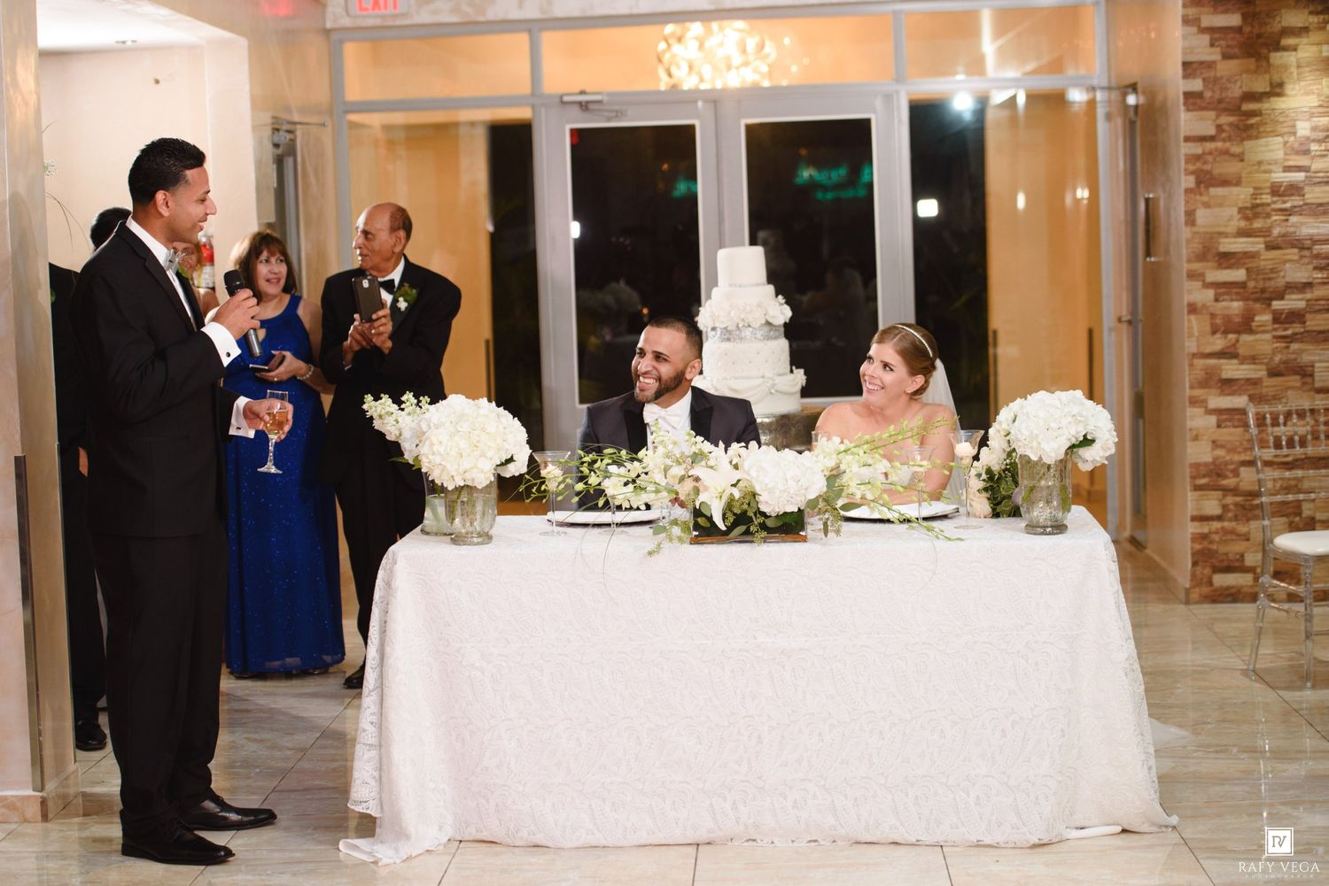 A man gives a toast at a wedding reception as the bride and groom smile at a decorated sweetheart table.
