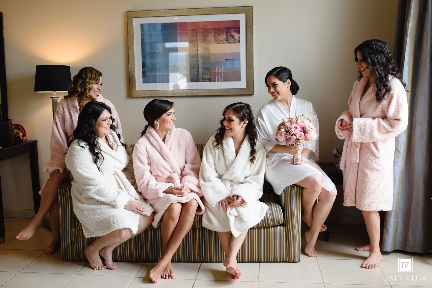 Bridesmaids in white and pink robes relaxing together in a hotel room before a wedding ceremony.