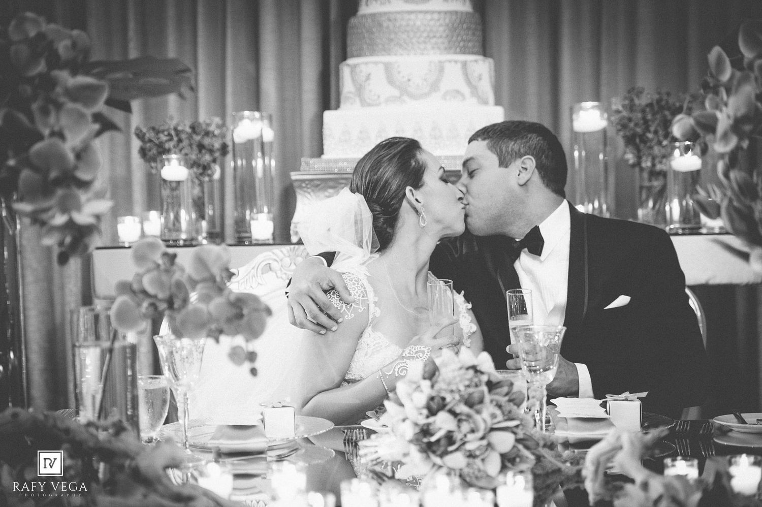 Black and white photo of a bride and groom sharing a kiss at their wedding reception table with an elegant cake backdrop.