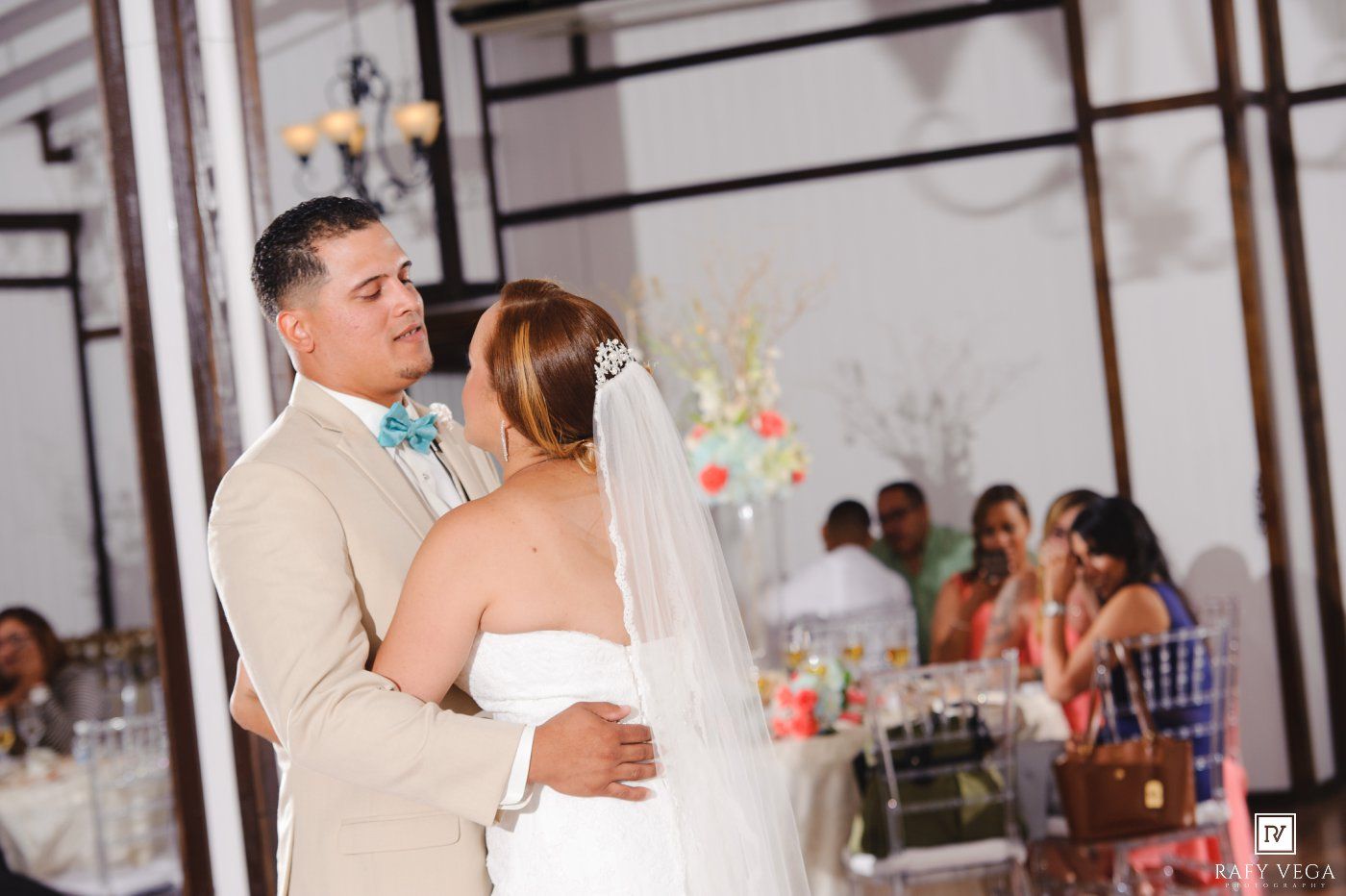 Bride and groom share their first dance at an elegant wedding reception with guests seated at decorated tables in the background.